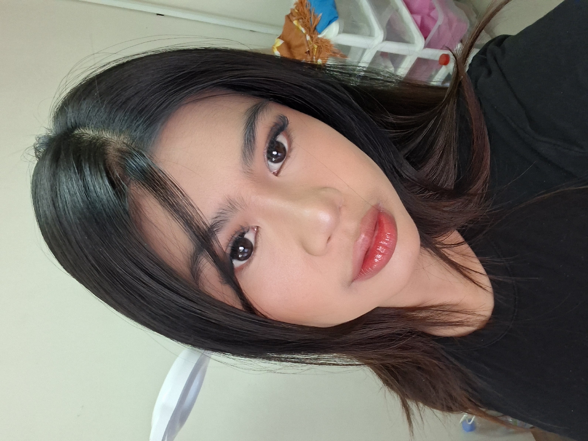 A young woman with long, dark hair and fair skin is photographed from the chest up. She is wearing a black t-shirt and has noticeable makeup, including red lipstick and false eyelashes. Her right eye is looking directly at the camera, while her left eye is slightly averted. The background appears to be an indoor setting, with a wall and a glimpse of storage containers with some items inside, possibly in Kuta, Indonesia. The lighting suggests it might be a personal space, such as a bedroom or dressing area.
