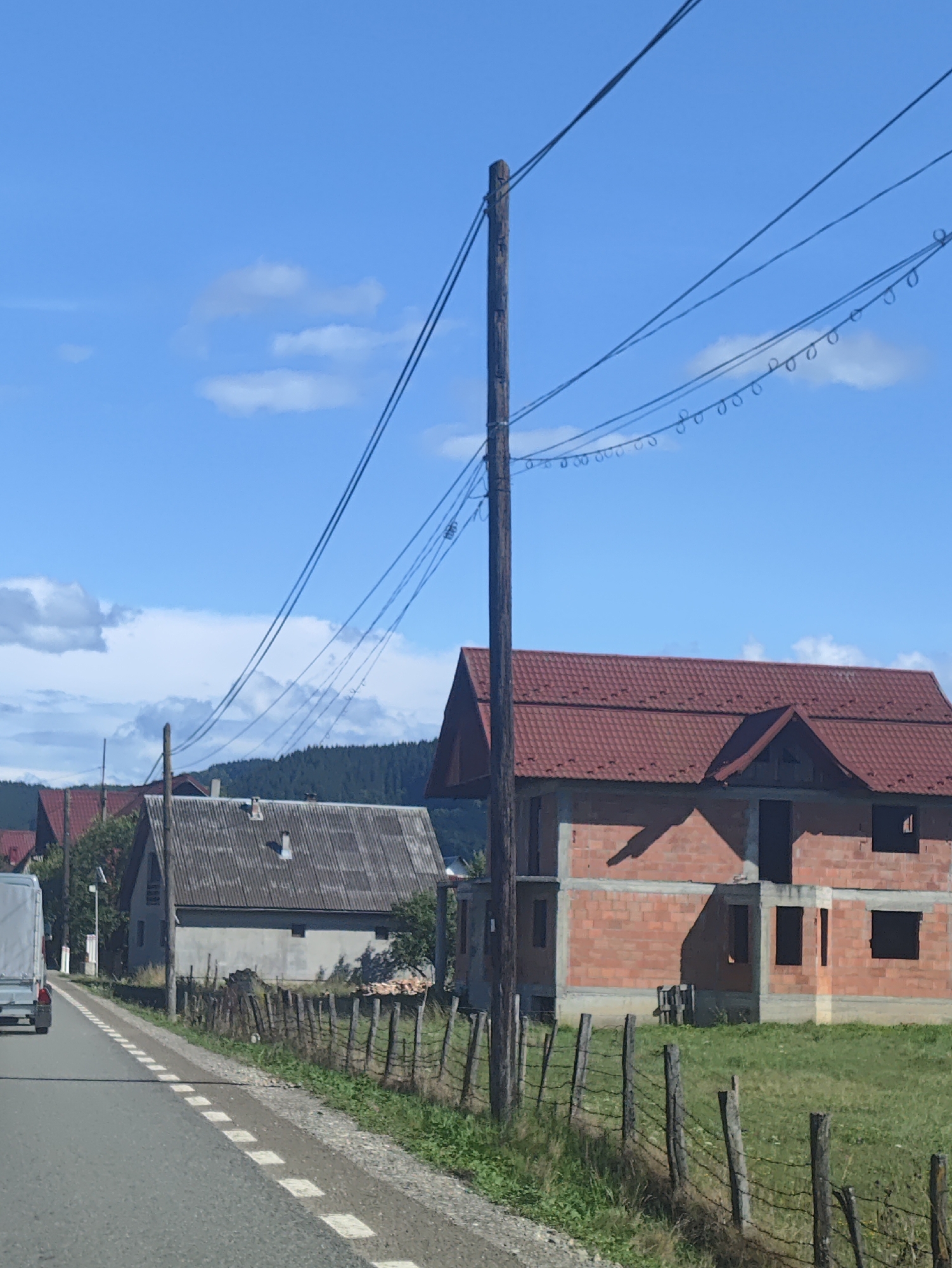 The image depicts a rural scene in Petrova, Romania, under a bright, partly cloudy blue sky, suggesting it is daytime and the weather is fair. The foreground shows a paved road with white dashed lines, bordered by a grassy verge and a rustic wooden fence.
Several houses are visible. To the left, a light-colored, older house with a grey, corrugated roof stands next to the road. Further down the road, a white truck is partially visible. To the right of the older house, a new, unfinished house constructed from red brick is prominent. It features a red tiled roof and has dark, empty window openings. Behind these structures, a dense line of evergreen trees forms a backdrop, indicating a forested area.
Overhead, a wooden utility pole supports multiple power lines, some of which are adorned with small, circular insulators. The overall impression is one of a developing rural community, with older dwellings alongside new construction. There is no visible text in the image.