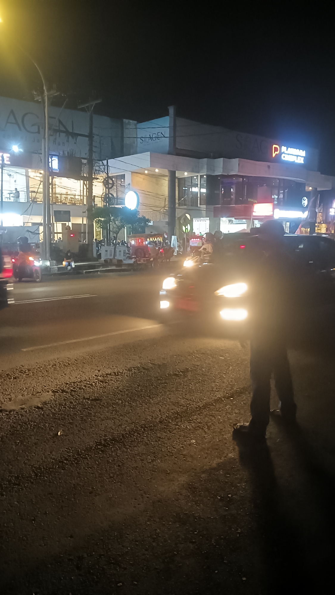 This is a night-time image taken from a low angle on a paved road in Sragen, Indonesia. The scene depicts a street with several vehicles, including cars and motorcycles, and a pedestrian on the right side of the frame. In the background, there is a multi-story building with illuminated signage, including "S-AGEN" and "PLATINUM COMPLEX." The street is wet, reflecting the lights of the vehicles and buildings. There are streetlights casting a yellowish glow. Several people are visible in the distance, some on motorcycles and others standing or walking. The pedestrian in the foreground is silhouetted by the streetlights, with their legs and feet visible, casting a long shadow on the ground. The overall impression is of a busy urban street at night.