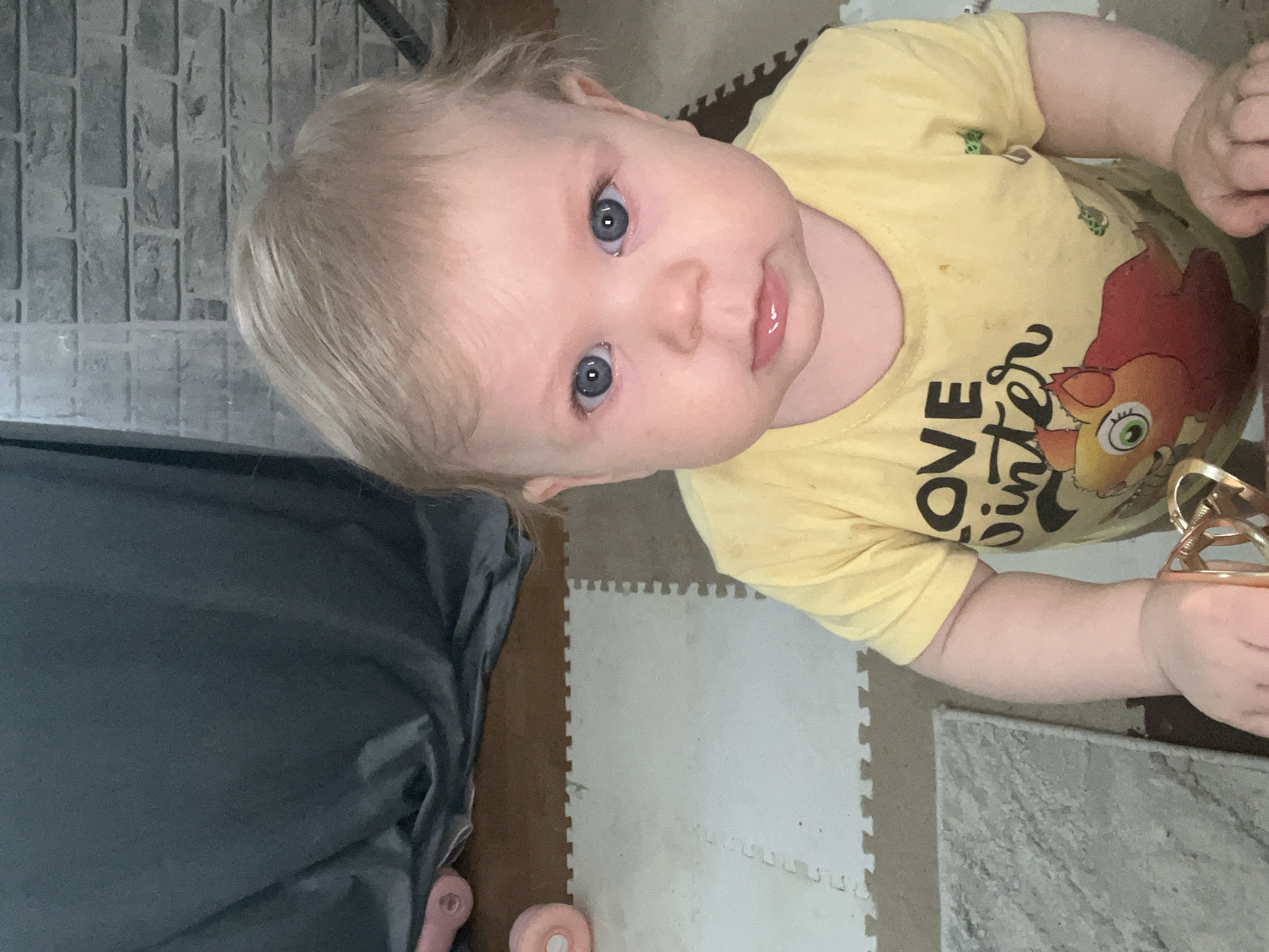 A close-up, tilted, indoor photograph shows a baby with blond hair and blue eyes, wearing a yellow t-shirt with a cartoon graphic on it. The baby is looking up at the camera. The background includes gray faux brick, interlocking foam floor tiles in white, brown, and gray, a dark gray, draped object (possibly furniture covered with a sheet), and a pink and orange object on the floor. The location is not possible to identify from the content in the image.