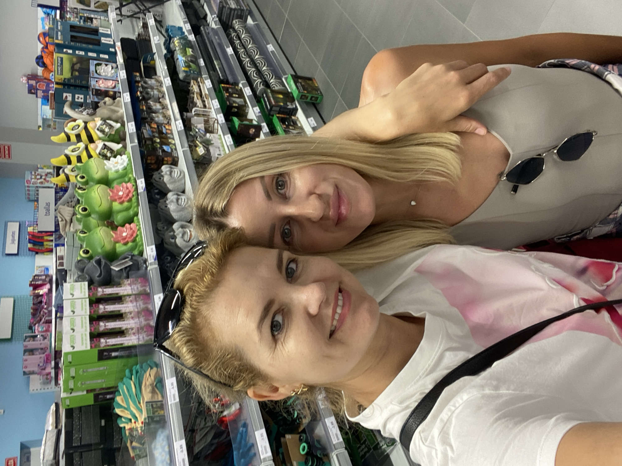 Two women are taking a selfie in a store. The woman on the left has blonde hair and is wearing a white t-shirt with a pink design. The woman on the right also has blonde hair and is wearing a grey tank top. They are standing in an aisle with shelves stocked with various items, including gardening supplies and decorative items. The background shows a blue wall and ceiling, and grey tiled flooring. The overall impression is that of a casual shopping trip in a retail environment. The image does not contain any specific elements that place it in the city of València, Spain.