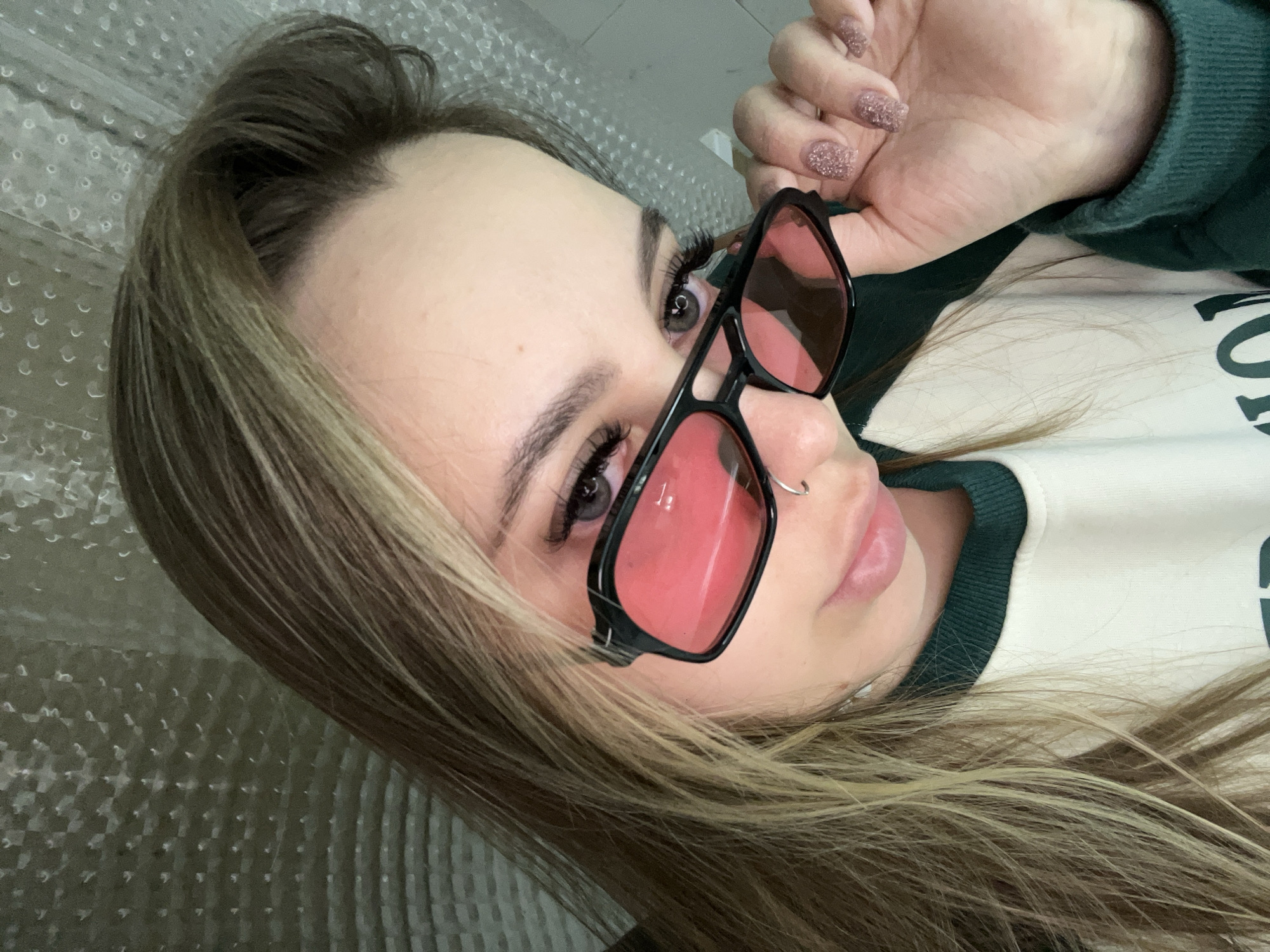 A young woman with light brown and blonde hair holds red-tinted sunglasses in front of her face. She has fair skin, dark eyebrows, and what appears to be a nose piercing. She is wearing a sweatshirt with green trim. The background appears to be a bubble wrap surface. The location is not discernible from the image.