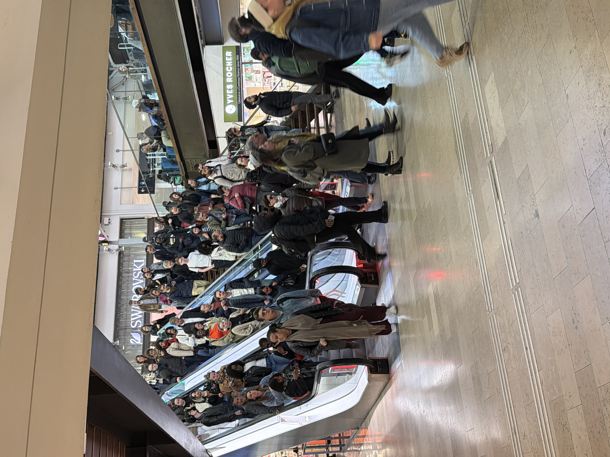 The image shows a busy scene in a shopping mall in Paris, France. A large crowd of people is riding an escalator. Stores, including Yves Rocher and Swarovski, are visible in the background. The floor is tiled. People are wearing winter clothing.