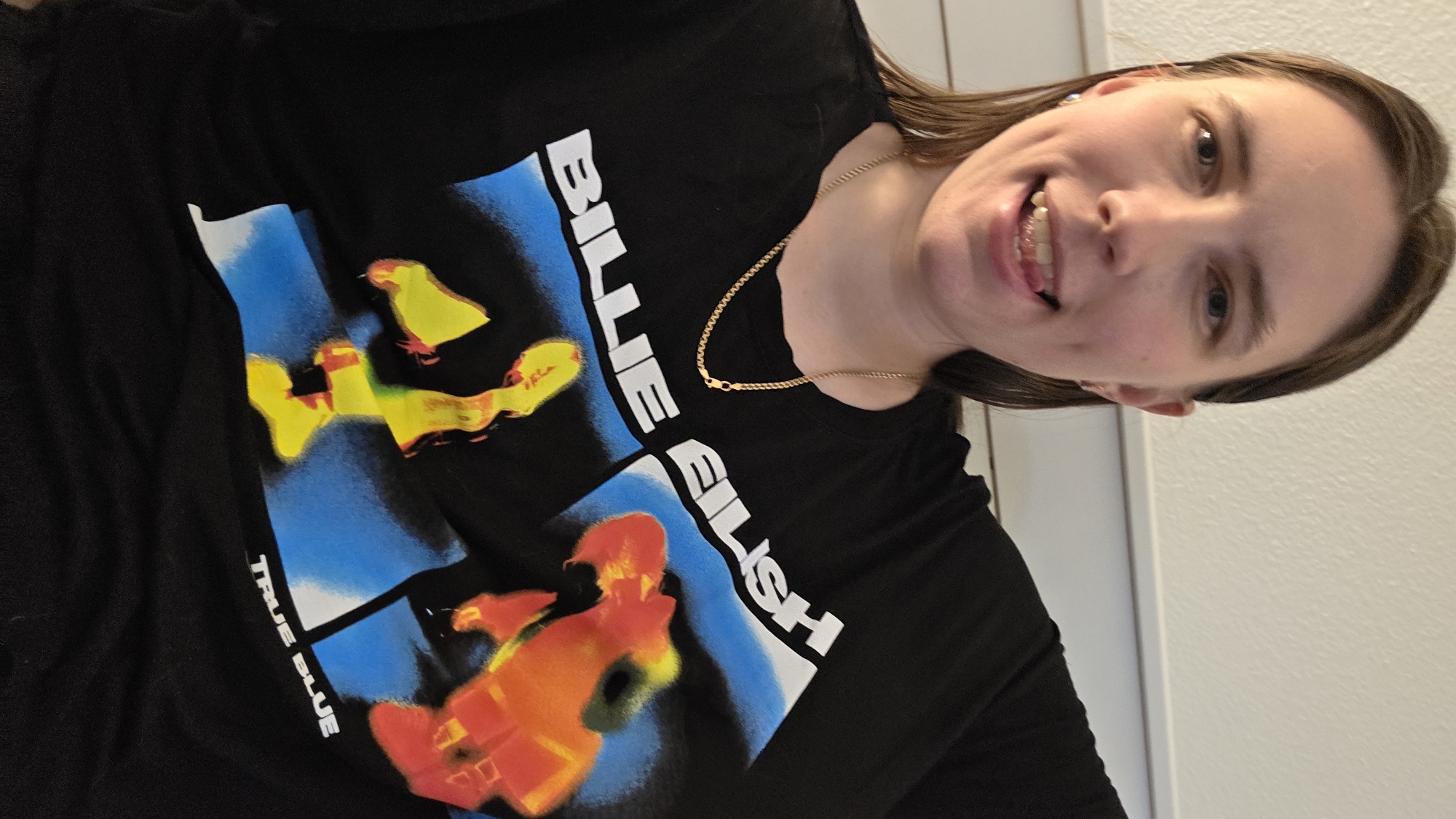 The image shows a medium shot of a woman wearing a black t-shirt with a graphic print. The print features the text "BILLIE EILISH" in white letters across the top, with abstract art below. The art consists of blurred figures in yellow and orange against a blue background. The woman is looking at the camera, with her tongue slightly sticking out. She is wearing a gold chain necklace and small earrings. The background is a plain, lightly textured wall. The image appears to be a selfie taken indoors, likely in a residential setting in Portland, United States of America.