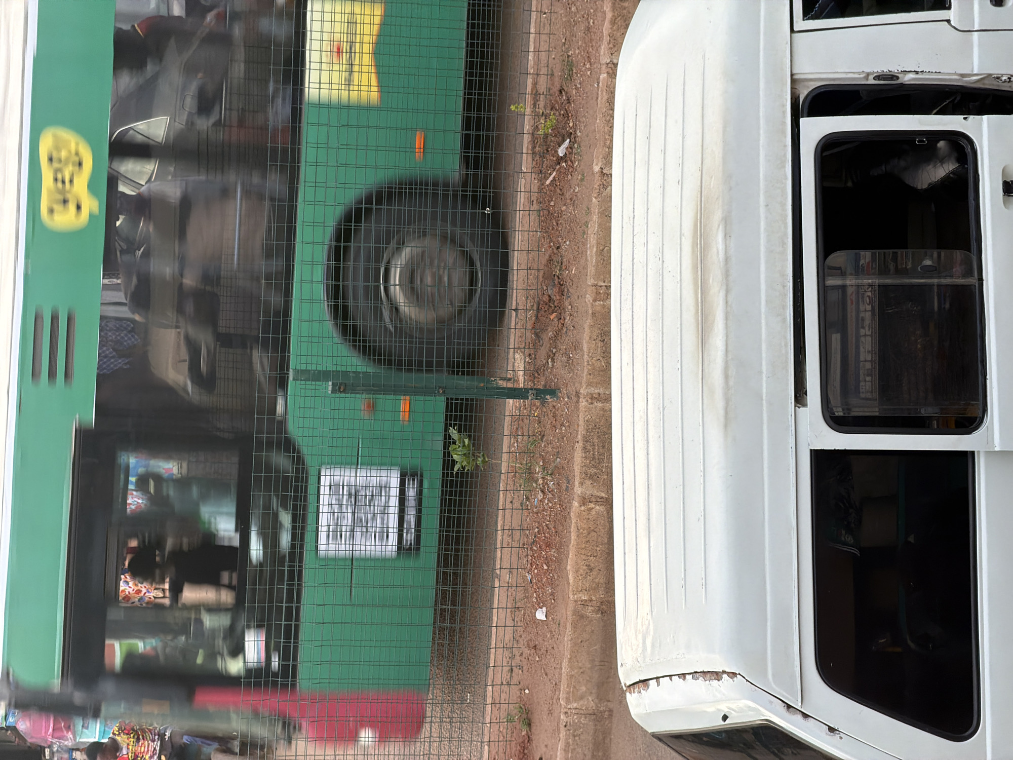 The image shows two vehicles parked next to each other on a dirt or gravel surface. On the left is a green bus with a yellow, cartoon-like graphic on its side. The bus has tinted windows and a wire fence in front of it. A large tire is visible behind the fence. The right side of the image shows the rear and side of a white van. The van has windows that appear to be tinted or covered from the inside. The rear of the van has a light-colored surface with visible vertical lines, and some rust is present near the bottom edge. The scene appears to be outdoors, with some sparse vegetation growing near the vehicles and fence. The lighting suggests it could be daytime. No specific activities are clearly depicted, but the vehicles are stationary. Visible text is limited to a white square sign with dark text on the green bus, and the yellow graphic on the bus, which contains some illegible text. The location is identified as Bukuru, Nigeria.