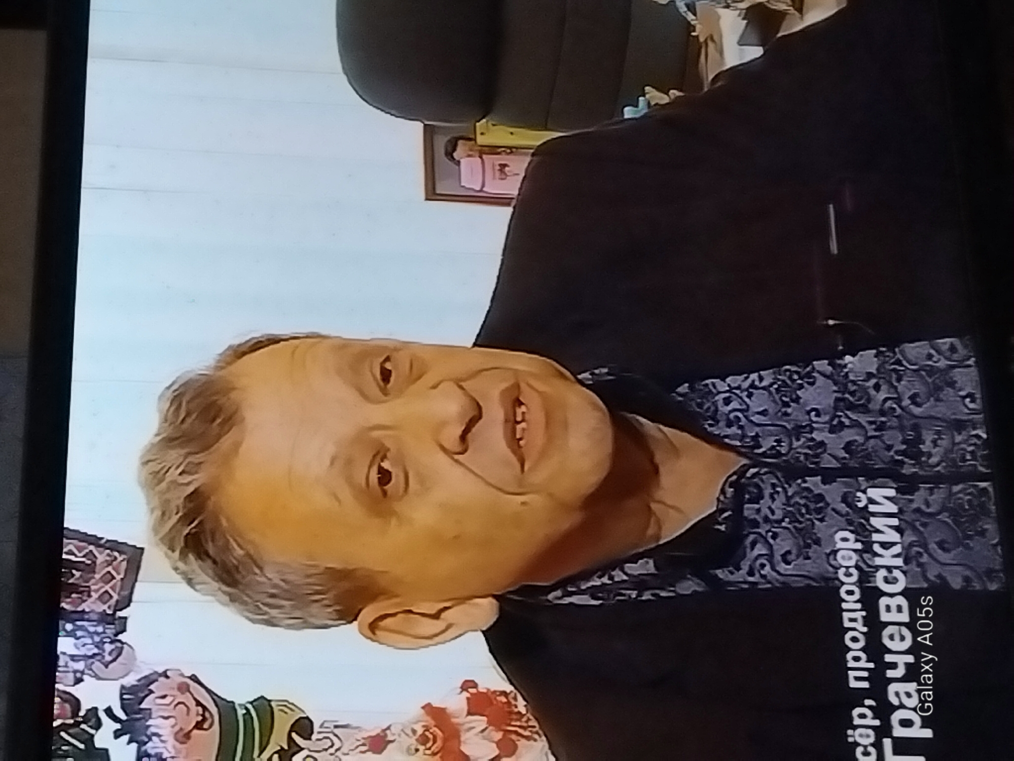 A close-up, medium shot shows a man with graying hair speaking, with a patterned shirt visible under a dark jacket. Text overlay in Russian reads "сёр, продюсер Грачевский" and below it "Galaxy A05s". To the left, partially visible, is a cartoonish figure. To the right, a black office chair and a framed picture are visible. There is no visible indication of the location in the city of Druzhba, Russia.