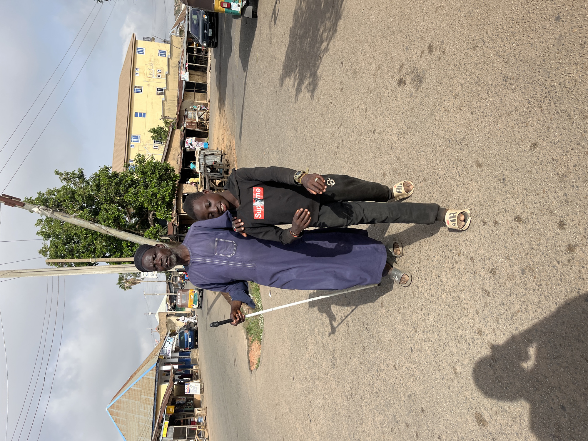 Two males stand on a paved road in Jos, Nigeria. One is an older man in a blue tunic and a black kufi cap, holding a white cane. The other is a younger man wearing a black "Supreme" hoodie. The younger man has his arm around the older man's shoulder. They both wear sandals. The background features buildings, trees, and overhead power lines. A vehicle is parked on the side of the road in the distance. The road surface appears worn.
