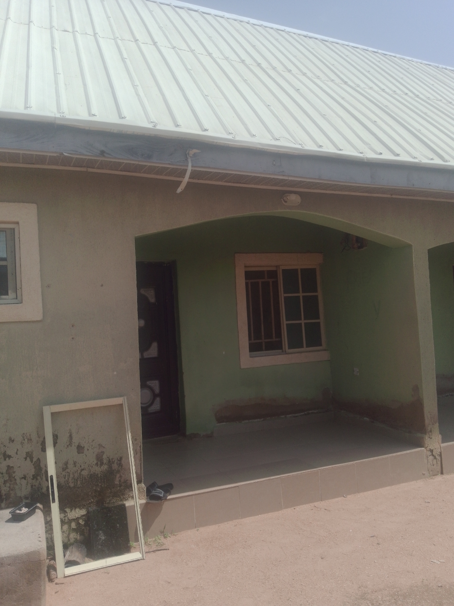The image shows the exterior of a single-story building with a light-colored metal roof. The walls of the building are painted a pale green. There is a covered porch area in front of the building. To the left of the porch, leaning against the wall, is a detached window screen. On the porch, a dark-colored door is visible, and a window with bars is set into the wall. Two pairs of black sandals are placed on the tiled floor of the porch. The ground in front of the building is made of sand and dirt. The scene appears to be in a residential area. Based on the context provided, this is the exterior of a house located in Jauro Yinu, Nigeria.