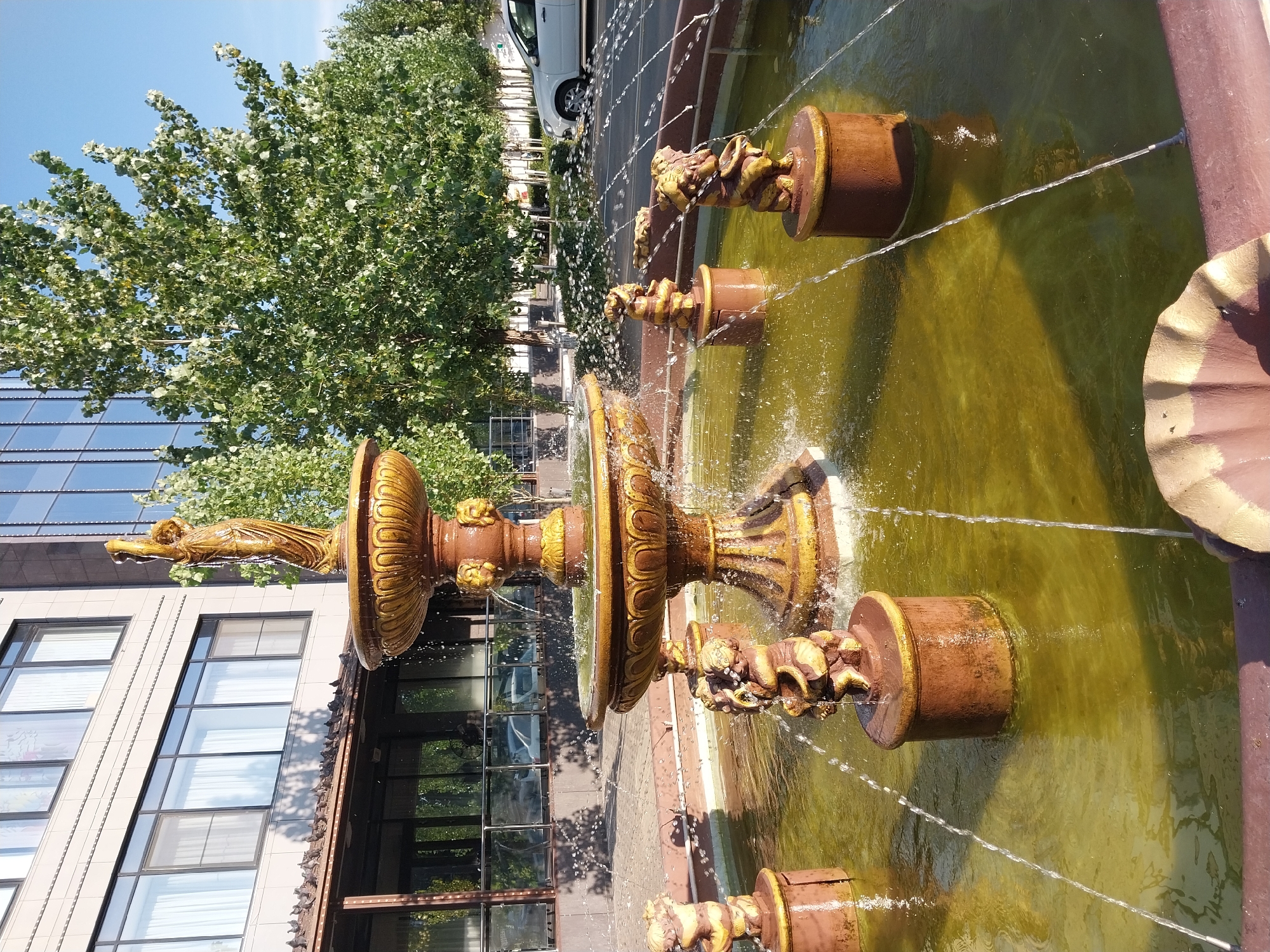 The image shows a fountain with multiple tiers and cherubic figures in a city park or public square. The fountain is made of a bronzed material, with intricate carvings and decorative elements. Water sprays from the fountain's various spouts, creating a dynamic visual effect. In the background, a modern building with glass windows and trees are visible, suggesting an urban setting. The sky is clear and blue, indicating a sunny day. The water in the fountain appears greenish, possibly due to algae or the color of the basin. The overall impression is one of a well-maintained public space, likely a popular spot for recreation and relaxation.