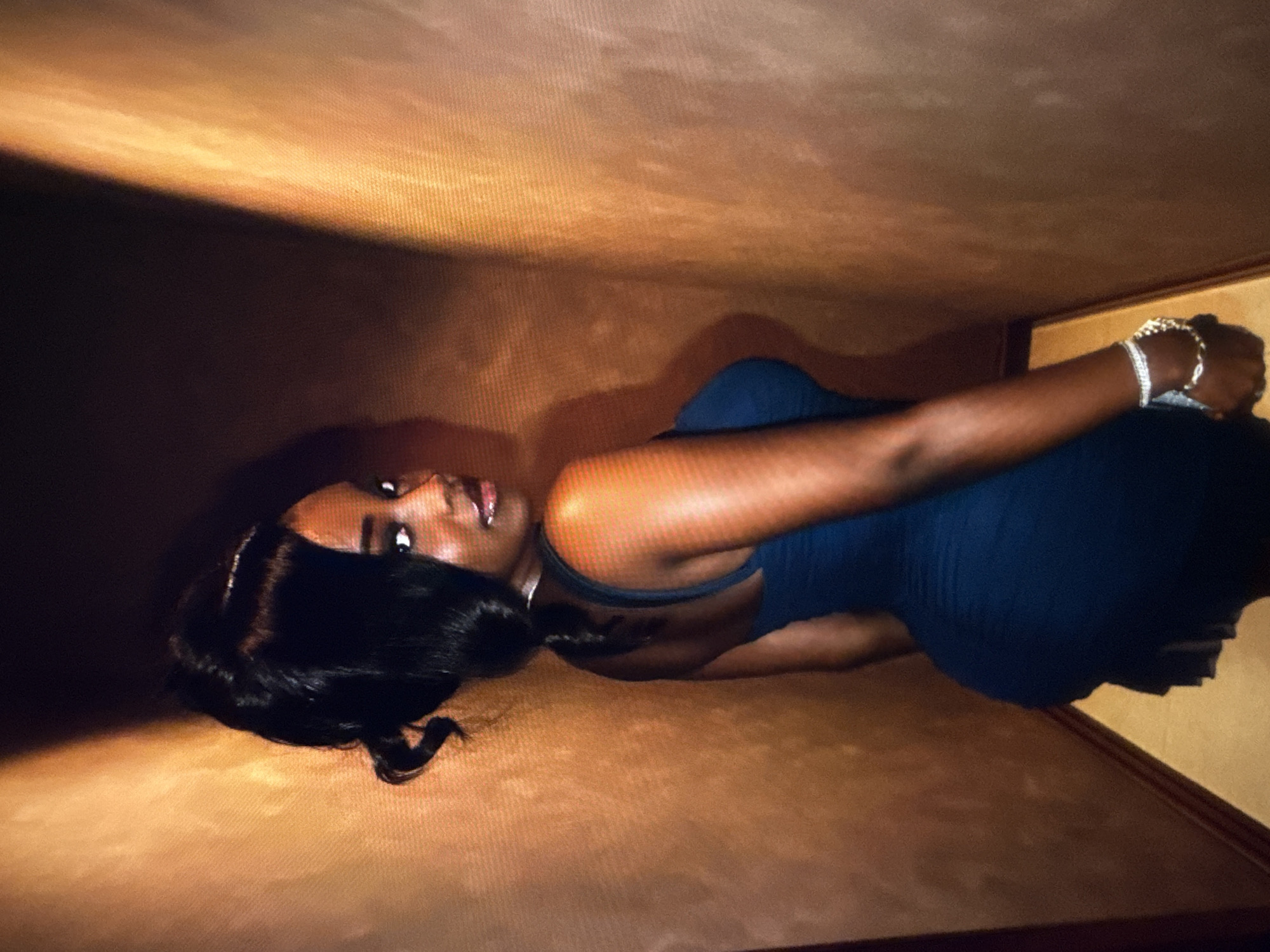 The image shows a Black woman with her back to the camera, looking over her left shoulder. She has dark hair styled in an updo with some loose curls. She is wearing a dark blue, form-fitting dress and a diamond bracelet on her left wrist. The background appears to be a narrow hallway with textured walls that have a gradient of light and dark brown tones. The lighting in the scene is warm and appears to be coming from the right, casting shadows on the left wall. There is no indication of the specific location within the city of Abuja, Nigeria.