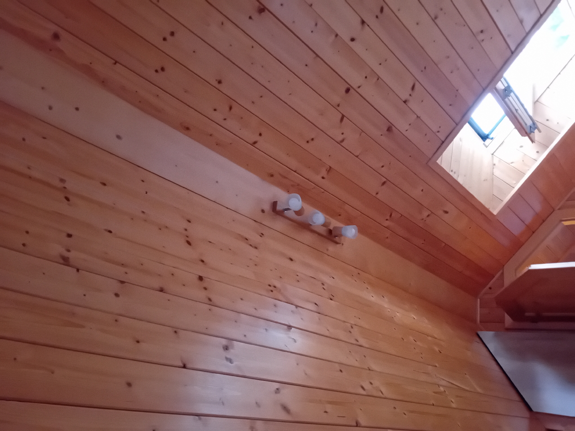 The image shows an interior view of a room with a sloped, wood-paneled ceiling. A row of three ceiling lights with white, frosted glass shades is mounted on the wooden planks. In the upper right corner, a skylight is visible, allowing natural light to enter the room. The wood grain and knots are clearly discernible on the ceiling. The lower left portion of the image shows a corner of what appears to be a dark, rectangular object, possibly furniture or a structural element, and a section of wooden doorway. The overall scene suggests a rustic or cabin-like interior. The location provided is Zuberec, Slovakia.