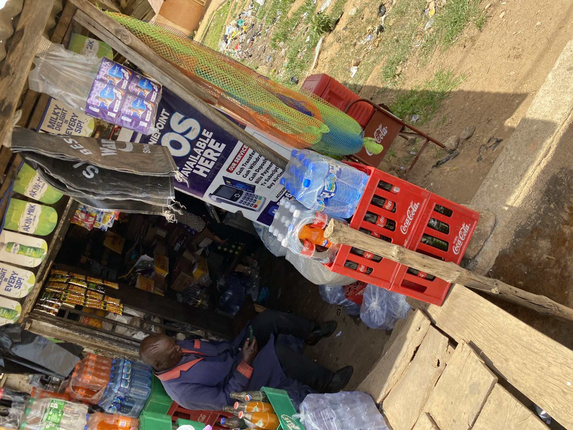 The image captures a street scene in Bukuru, Nigeria, showing a small, open-front shop crammed with merchandise. A man in a blue jacket and dark trousers is sitting on the ground inside the shop, holding a cell phone. The shop's inventory includes bottled water, soft drinks in crates (Coca-Cola and Fanta), milk products ("Milky Delight," "Aktiv-Milk"), and packaged goods. An advertising sign displays the services available, such as "Cash Transfer," "Cash Withdrawal," and "Bills Payment." Outside the shop, there's a Coca-Cola cooler on a small metal stand. The surrounding area shows signs of litter and vegetation. The shop appears to be constructed from wooden planks and corrugated metal.