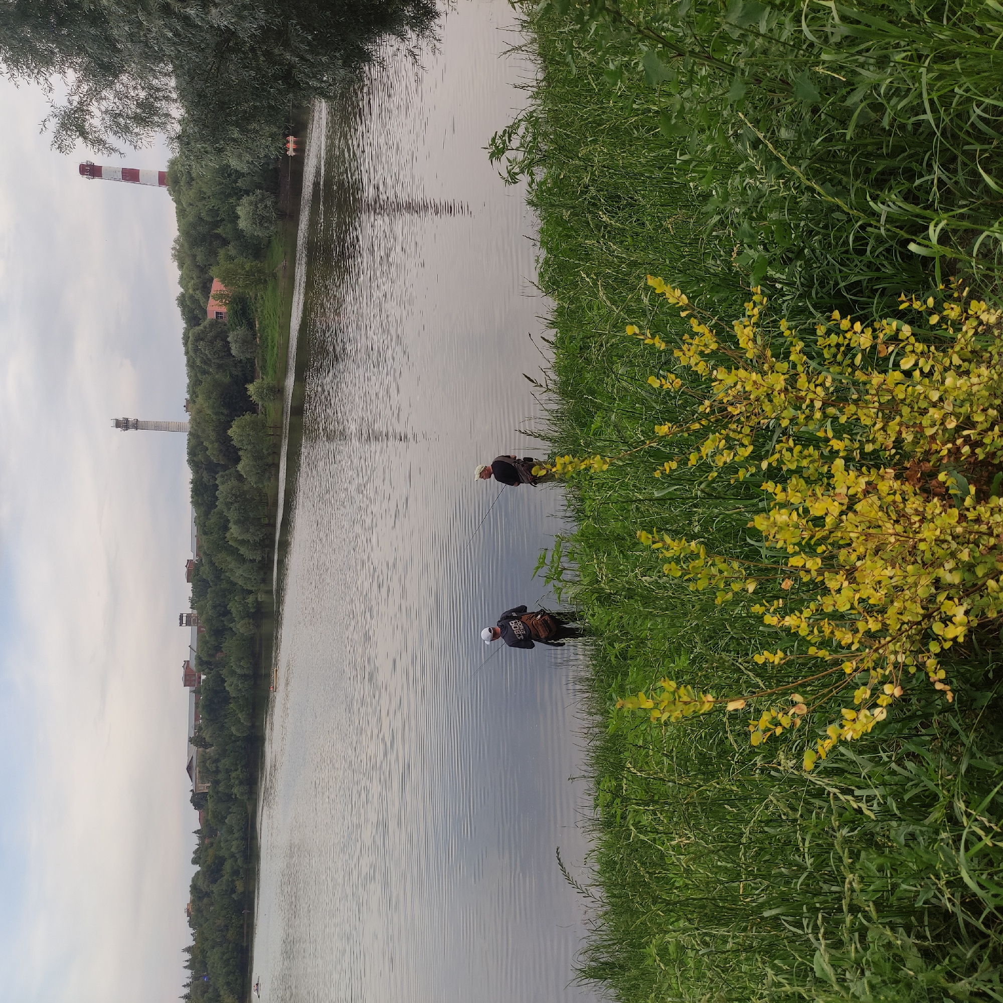Two men are fishing in a wide, calm river with rippling water. They are standing on the grassy bank in the foreground. The man on the left is wearing a white cap and a dark hoodie with a backpack slung over his shoulder. The man on the right is wearing a light-colored cap and a dark shirt, also with a backpack. Behind them, across the river, is a dense line of trees separating the water from a flat landscape with buildings and industrial structures, including a tall, red and white striped smokestack and a white cellular tower. The sky is overcast and the overall lighting suggests it is late afternoon or early evening. The foreground is dominated by tall green grass and a bush with yellow leaves.