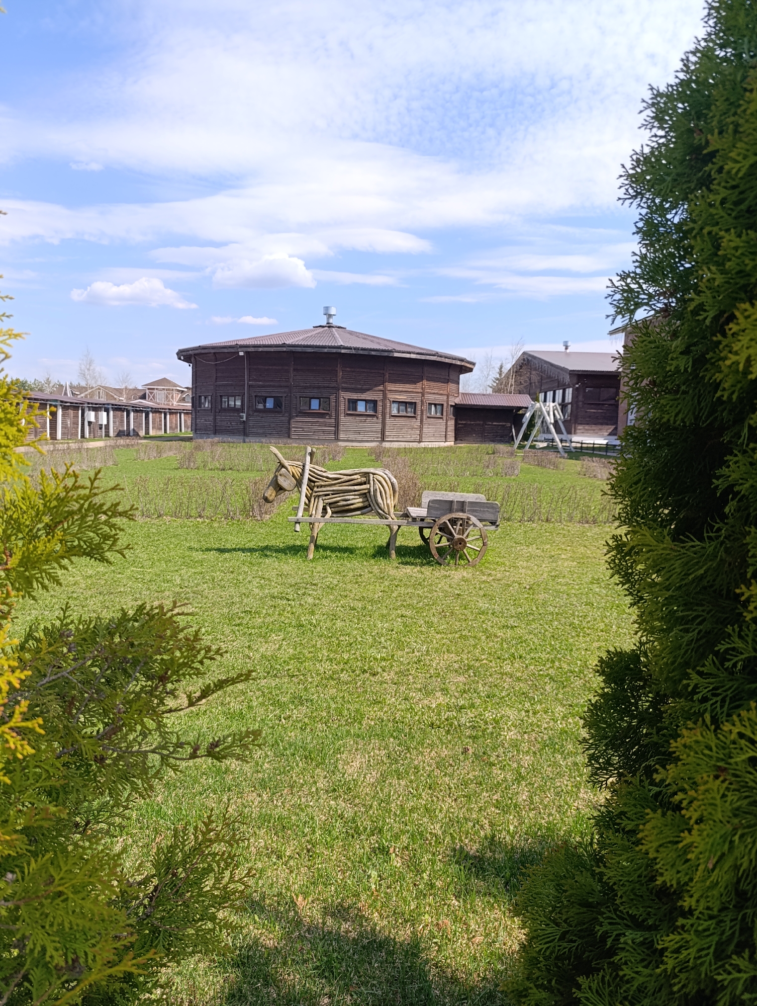 The image shows a grassy lawn featuring a wooden sculpture of a donkey pulling a cart, with a large, round wooden building in the background. The scene is framed by greenery on either side.
The donkey sculpture is made of branches and twigs, with a head, body, and legs formed from the wood. The cart is also made of wood, with four wheels and a flat top.
The round wooden building in the background has a brown roof and multiple windows. Other wooden structures are visible to the left and right of the main building.
The lawn is green and well-maintained. The sky is blue with white clouds.
