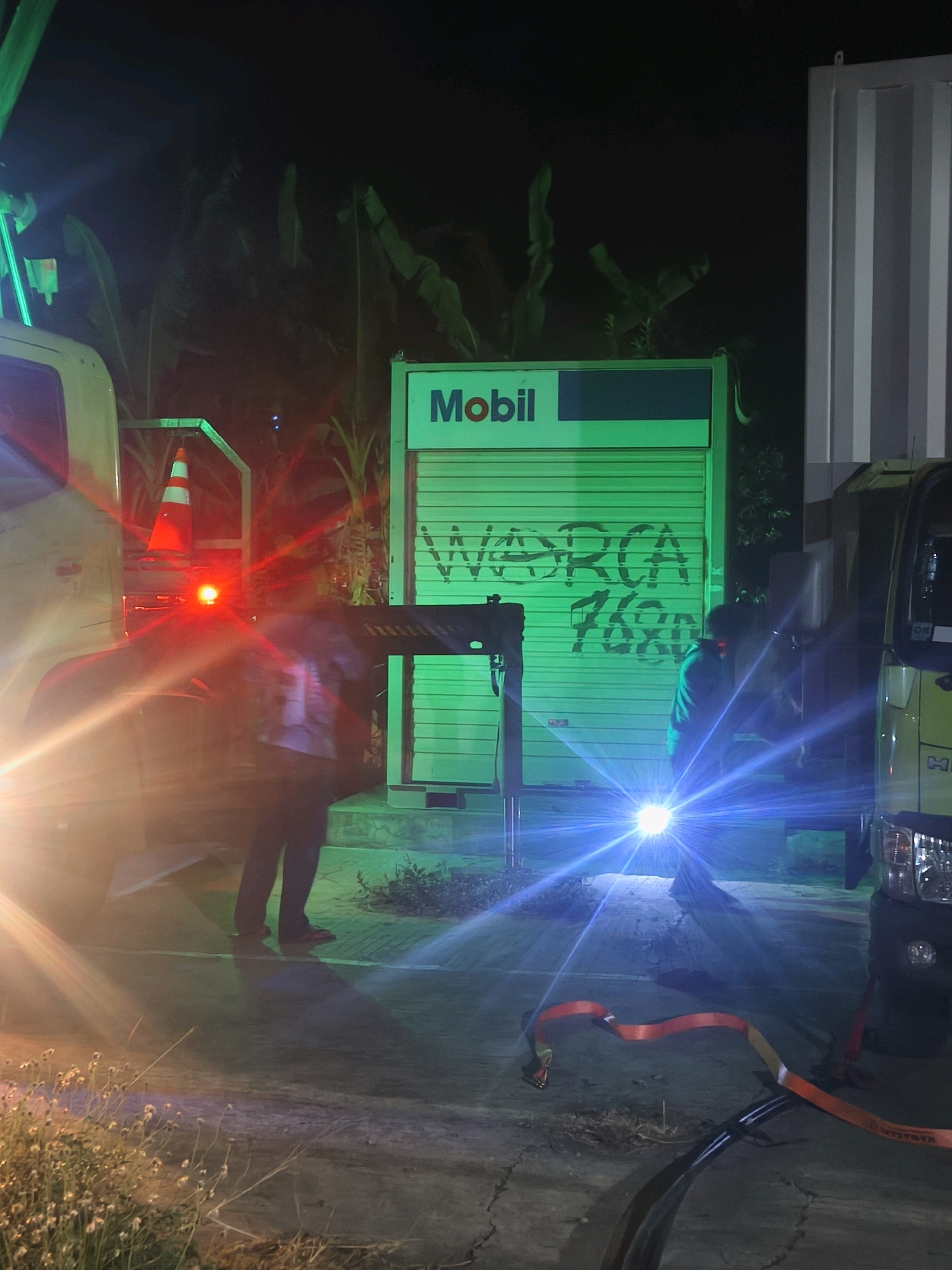At night, a Mobil branded container, illuminated by green lights, is visible in what appears to be a service station or storage area in the city of Kroya, Indonesia. The container's metal roller shutter door is marked with graffiti. Two vehicles, a light-colored truck on the left and a darker truck on the right, are present. One individual, wearing dark pants and a light-colored shirt, stands near the container. Another individual, wearing dark clothing, is partially obscured by a bright blue light emanating from a portable light source. The ground is paved, with some debris and dry grass visible. An orange strap lies on the ground. The scene suggests an ongoing operation or activity at the location.