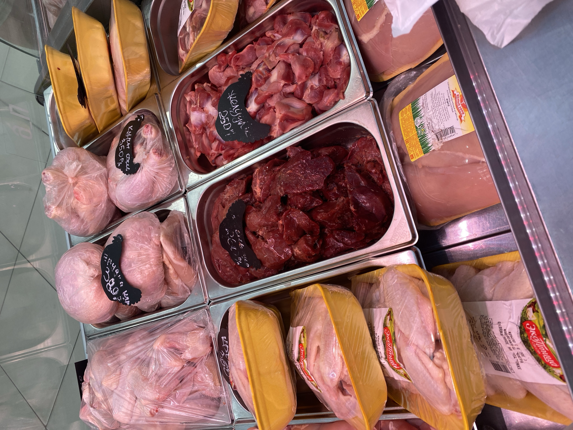 This media file captures a close-up, overhead view of a refrigerated display case filled with various raw poultry products, likely at a market or grocery store in Novaya Usman', Russia. The focus is on the different cuts of chicken and possibly other fowl, presented in clear plastic packaging and metal trays.
In the upper section, several trays hold what appears to be chicken gizzards and liver. The gizzards are a pale pinkish-red, while the liver is a darker, rich red. Some of these are marked with handwritten labels, one indicating "желудк 250" and another with "куры сocel 350", suggesting the type of meat and price. To the left, there are three large, oblong packages with golden yellow bases, possibly containing larger cuts of meat like whole chickens or turkey parts, wrapped in clear plastic.
Moving down, there are multiple packages of chicken parts. Some are whole chickens or large portions presented in yellow trays, with labels that include "ЯСНЫЕ ЗОРИ" and "Эко Птица," indicating the brand and product type like "крыла" (wings). Other chicken pieces, such as wings and what look like drumsticks, are visible in clear plastic bags. One bag has a handwritten label with "Ayam KAP 38 0".
The overall setting appears to be a commercial food display. The lighting is bright and artificial, typical of a refrigerated retail environment. The image is sharp, highlighting the textures and colors of the raw meat. There are no people visible, and the scene is focused solely on the products for sale. The visible text, predominantly in Russian, provides brand names, product descriptions, and pricing information. The overall impression is one of a well-stocked and organized meat counter.