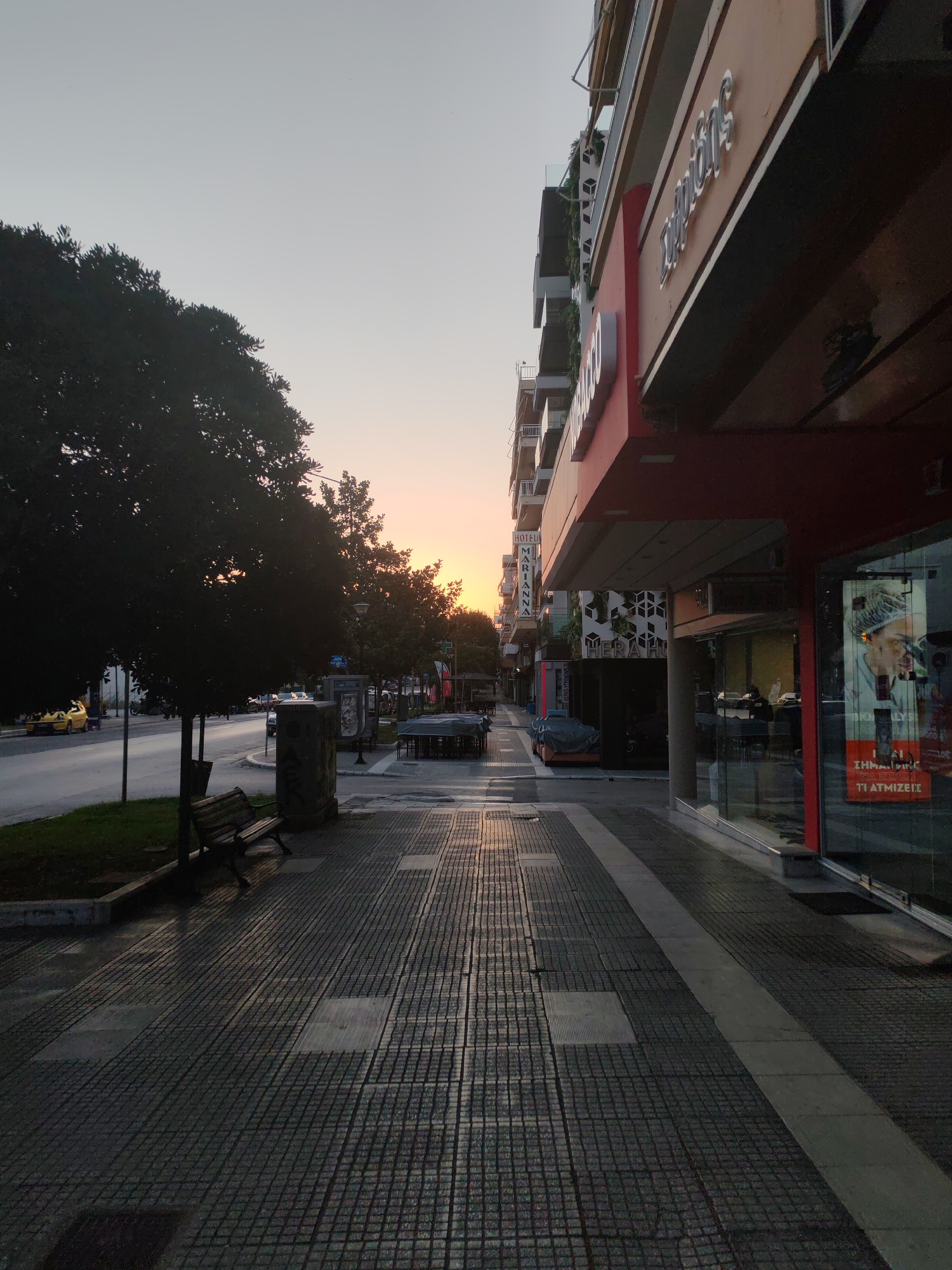 The image depicts a street scene in Alexandroupolis, Greece, during what appears to be sunrise or sunset. The perspective is from a sidewalk looking down a street lined with buildings on the right and trees and a street on the left.
On the right, a multi-story building is prominent, with a red-accented facade featuring signage that includes the word "ΣΤΕΦΑΝΟΣ" in large, silver, three-dimensional letters. Below this, a shop window displays an advertisement with text that includes "ΒΙΟΑΝΑΛΥΣΗ" and "ΤΙ ΑΤΜΙΖΕΙΣ". Another building further down the street has a vertical sign that reads "HOTEL MARIANNA".
On the left side of the street, a dark-leafed tree is silhouetted against the sky. A portion of the street is visible, with a yellow taxi and other vehicles in the distance. A sidewalk with a patterned tile surface runs along the foreground. A wooden bench is positioned on the sidewalk near a trash receptacle. In the distance, several outdoor seating areas with covered tables are visible, suggesting the presence of cafes or restaurants. The sky exhibits a gradient of light blues and oranges, indicative of the time of day. The overall atmosphere is calm and quiet.