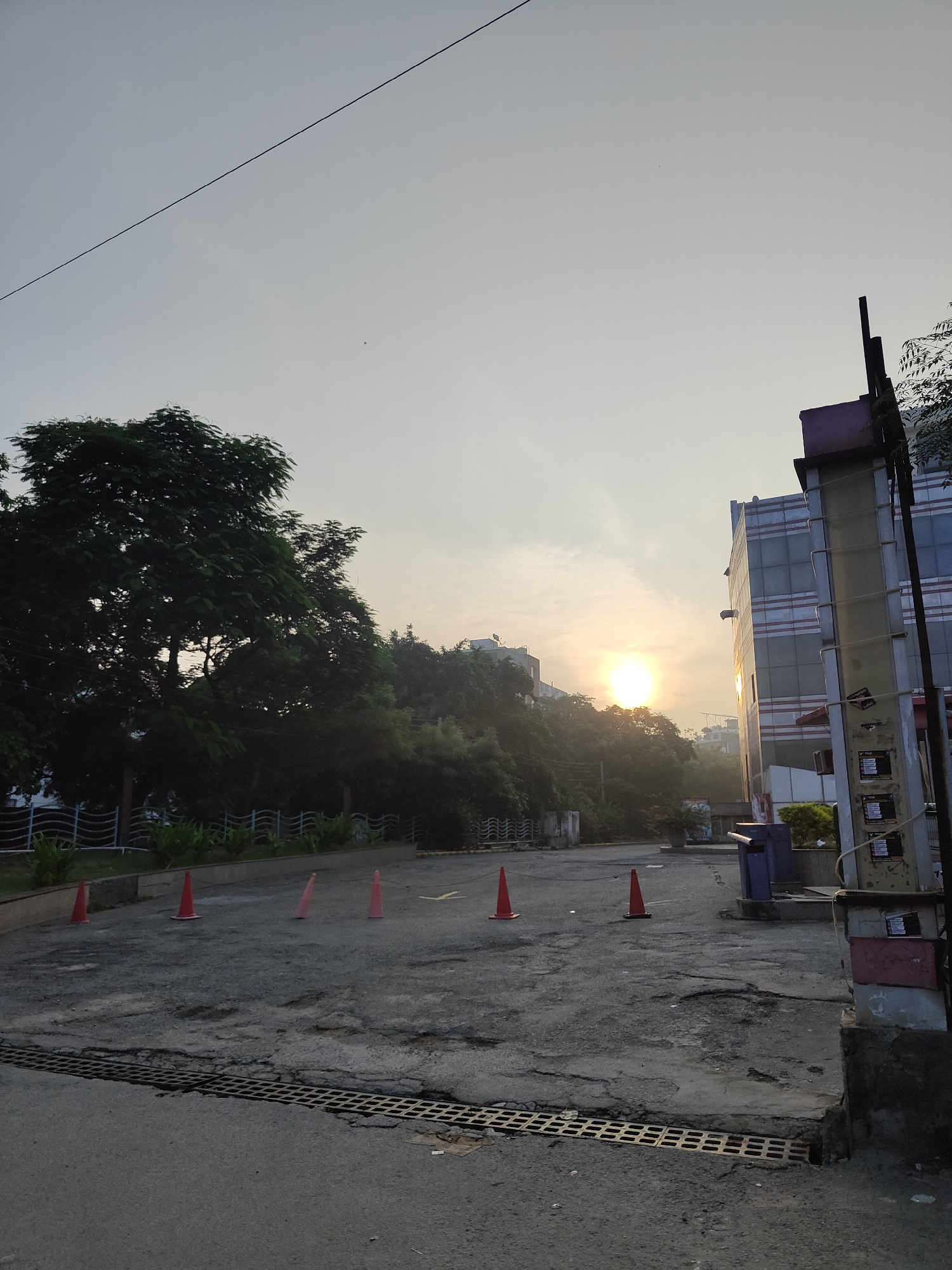 An outdoor, daytime, full-frame vertical shot captures a street scene in Gurgaon, India, during sunrise. The sky is a pale gradient of yellow and white, with the sun a bright, diffused orb peeking through a hazy atmosphere on the left side of the frame. A thin black power line bisects the upper portion of the sky diagonally.
In the middle ground, a paved area with visible potholes and cracks stretches across the foreground. Several orange traffic cones are scattered on the pavement. A white, wavy fence runs along the background, separating the paved area from a lush green slope covered in trees.
To the right, a modern, multi-story building with a reflective glass facade stands tall. The building's exterior features horizontal bands of red and grey. Adjacent to the building is a tall utility pole with various wires and mounted boxes. A blue trash bin is visible near the building entrance.
The overall impression is one of an urban environment bathed in the soft light of early morning, with a contrast between natural elements like trees and the man-made structures of the city. There are no people visible in the image.