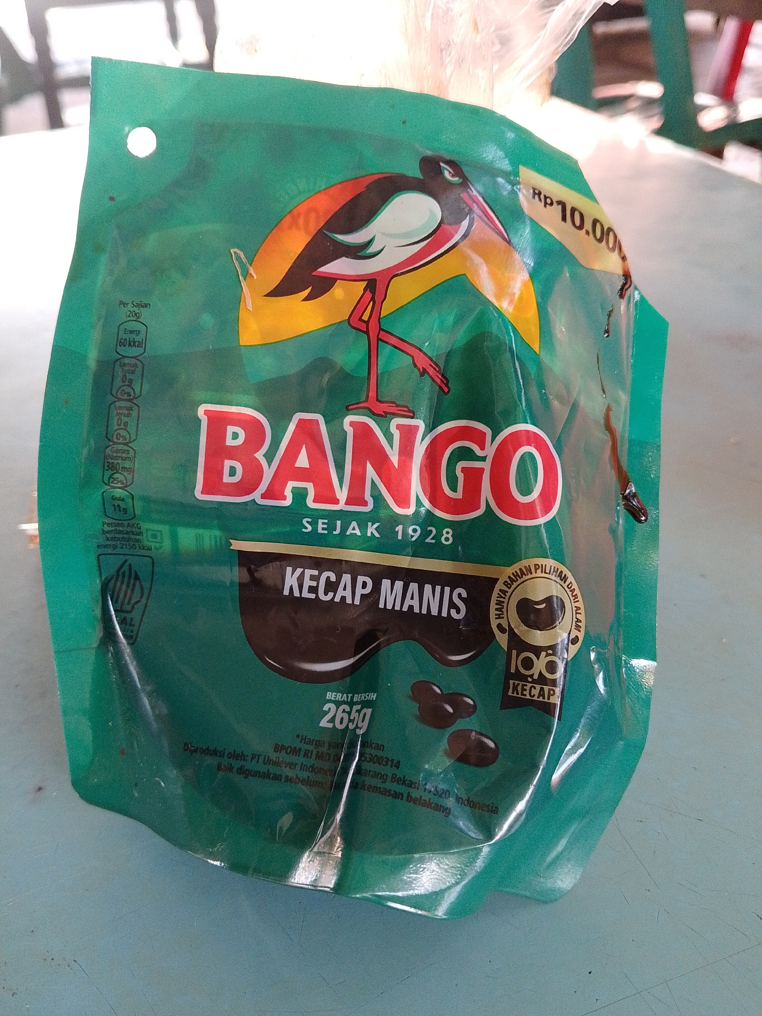The image shows a green plastic packet of Bango Kecap Manis (sweet soy sauce). The packet features a logo with a bird and the word "BANGO" in red letters. Below that, it says "SEJAK 1928" (since 1928). Below this is an image of a brown splash, likely representing the sauce. The words "KECAP MANIS" are printed on the brown splash. Underneath the brown splash there are three brown oval shapes, likely representing the soybeans used to make the soy sauce. The label indicates the product weighs 265g and has a price of Rp10.00. The packet also includes nutritional information and manufacturer details. It indicates that the product is made by PT Unilever Indonesia in Karang Bekasi 17520, Indonesia. There is no clear information to confirm the location of Lhokseumawe.