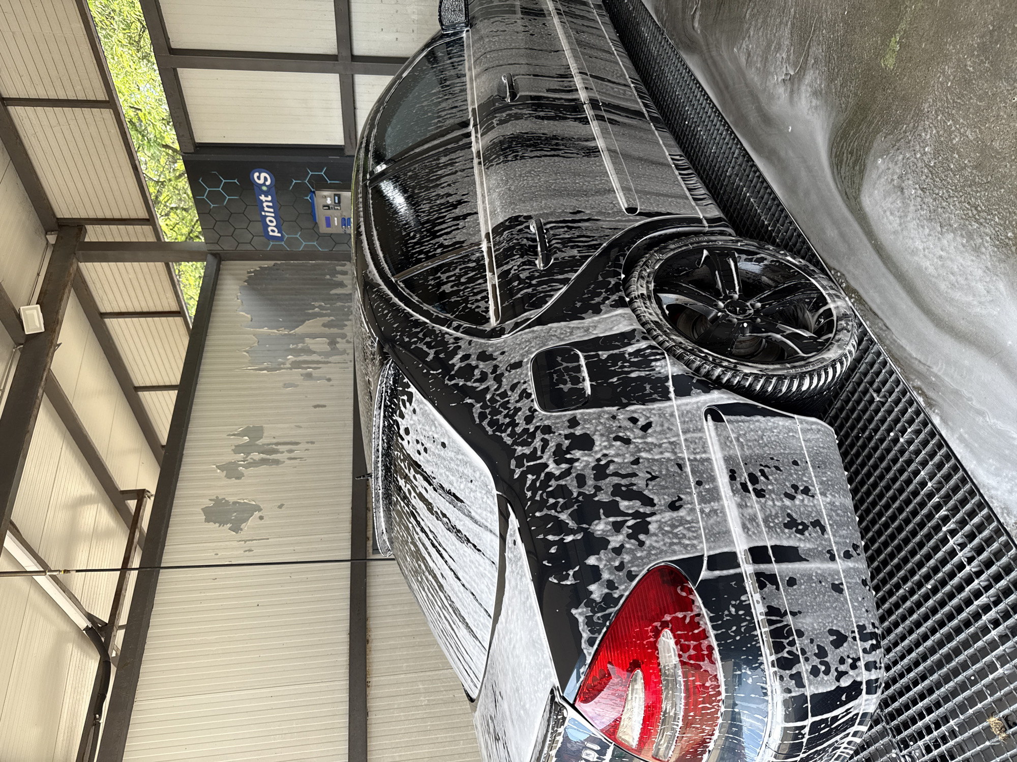A black Mercedes-Benz E-Class sedan (W211 generation) is captured mid-wash at a self-service car wash bay located in Stara Zagora, Bulgaria. The vehicle is thoroughly coated in white soap suds, which stream down its black body panels, rear window, and tinted side windows, with the left rear taillight illuminated. The right rear wheel features a black multi-spoke rim. The car is positioned on a black metal grate floor designed for drainage, which is visibly wet with suds and water. The surrounding car wash structure consists of white corrugated metal walls, some exhibiting water stains, and exposed metal support beams. In the background, a dark wall with a hexagonal pattern displays a "point S" branded payment console. Overhead, an opening in the structure reveals green tree foliage, indicating an outdoor or semi-outdoor setting. A slender, black radio antenna extends vertically from the vehicle's roof.