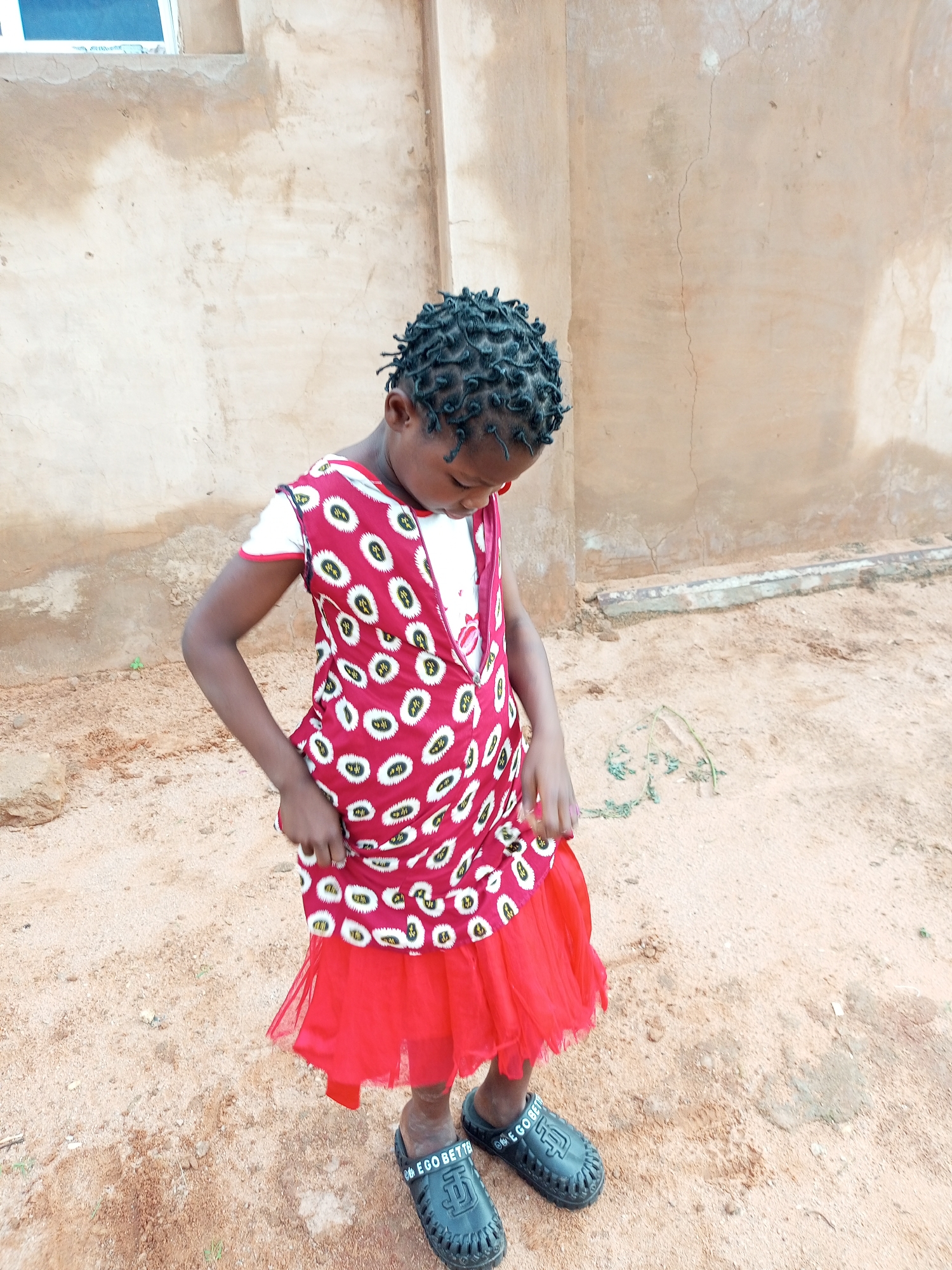 A young Black girl with braided hair, wearing a red and white patterned dress and a red tutu, stands on sandy ground in front of a beige, cracked wall. She is looking down, holding the fabric of her dress. She wears black slides with white text on them. A small, green plant is visible on the ground to her right. The scene appears to be outdoors in a rural or semi-urban setting.