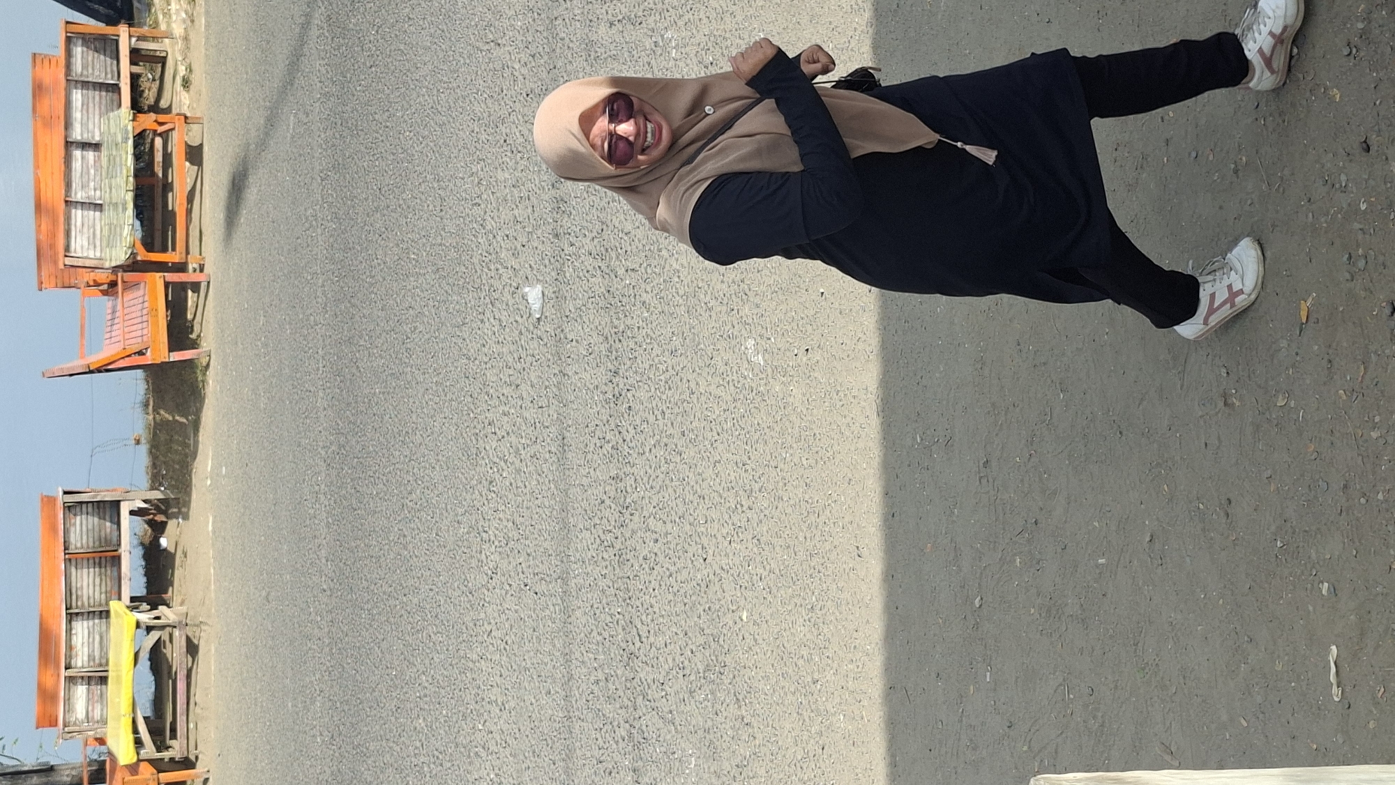 A woman wearing a black dress, black pants, and a light brown hijab is walking on a paved surface in Lhokseumawe, Indonesia. She is also wearing sunglasses and white sneakers with pink accents. The woman is smiling and appears to be in motion. In the background, there are several benches with orange and wooden frames situated on a raised area. The sky is visible above the benches, and the environment appears to be outdoors.