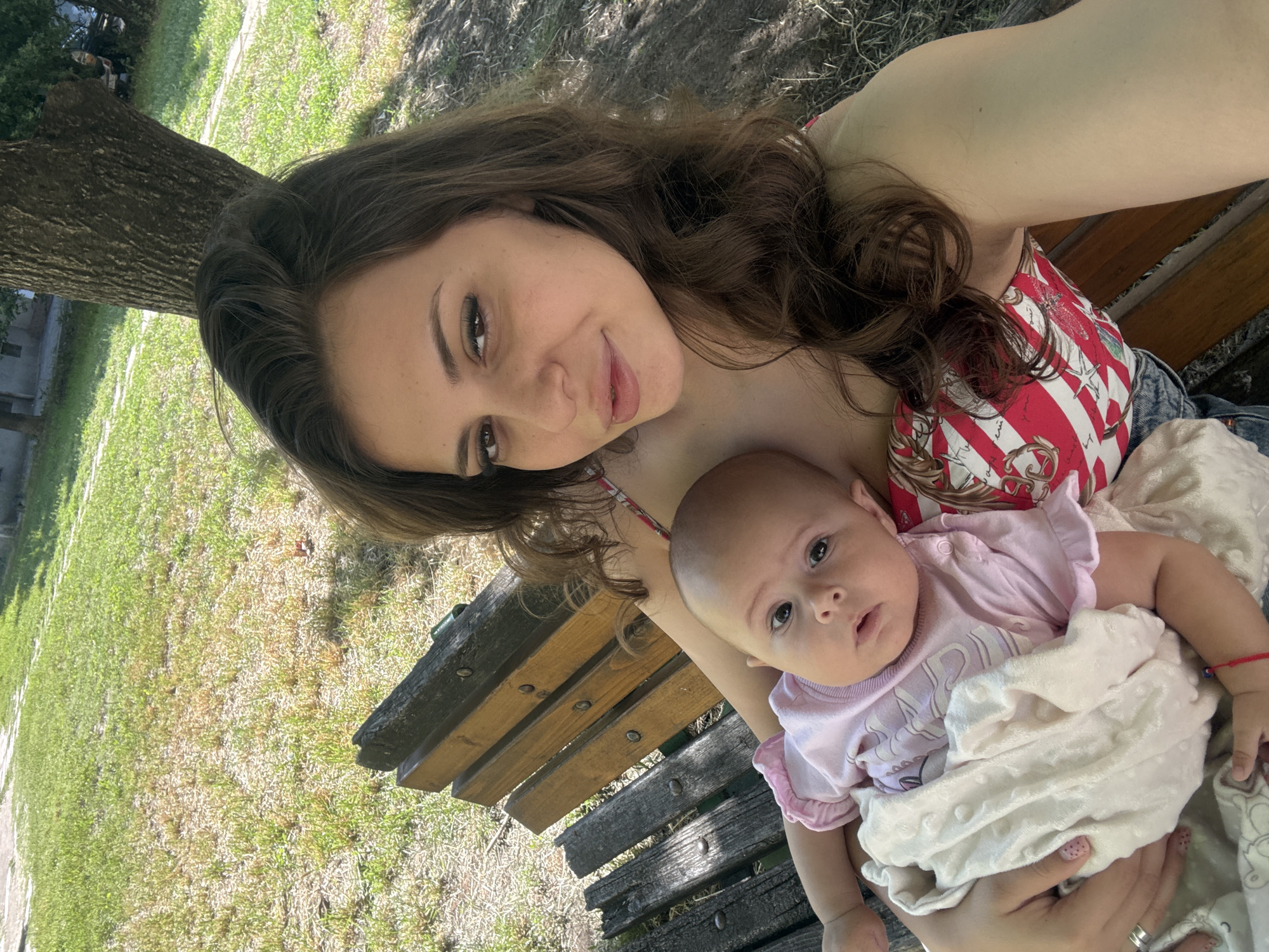 A young woman with long brown hair and light skin is holding a baby on her lap while sitting on a wooden park bench. The woman is wearing a red and white striped top and blue jeans. The baby is wearing a pink onesie. The background shows a grassy area with trees and a paved path. The scene appears to be in a park in the city of Plovdiv, Bulgaria, specifically in the Tsar Simeon's Garden, known for its mature trees and recreational spaces. The lighting suggests it is daytime, likely sunny.