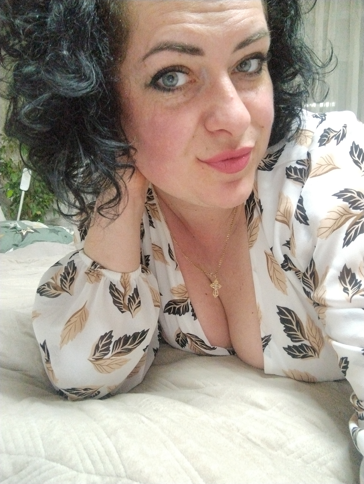 A woman with curly black hair and blue eyes is lying on a bed. She is wearing a white, long-sleeved shirt with a leaf pattern and a gold necklace with a cross pendant. Her left hand is resting on her neck. The background is blurred, but a plant and a curtain are visible. The location is not discernible from the image and cannot be identified as Grishanki, Russia.