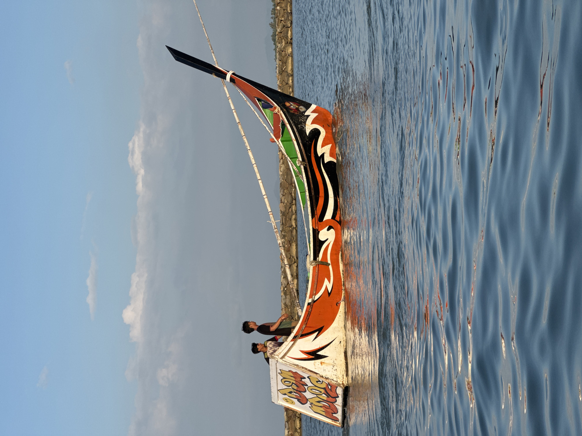 The image depicts a fishing boat on calm water, likely in Meunasah Jurong, Indonesia, under a bright, partly cloudy sky. The boat is vibrantly painted with orange, black, white, and green designs, featuring a striking tiger-like graphic on the hull and lettering on the stern. Two young men, dressed casually, are visible on the boat, one standing and observing the surroundings, the other looking forward. The water shows gentle ripples reflecting the light. In the background, a stone breakwater is visible, and beyond that, a distant shoreline with vegetation. The scene captures a moment of stillness during what appears to be a calm day on the water.