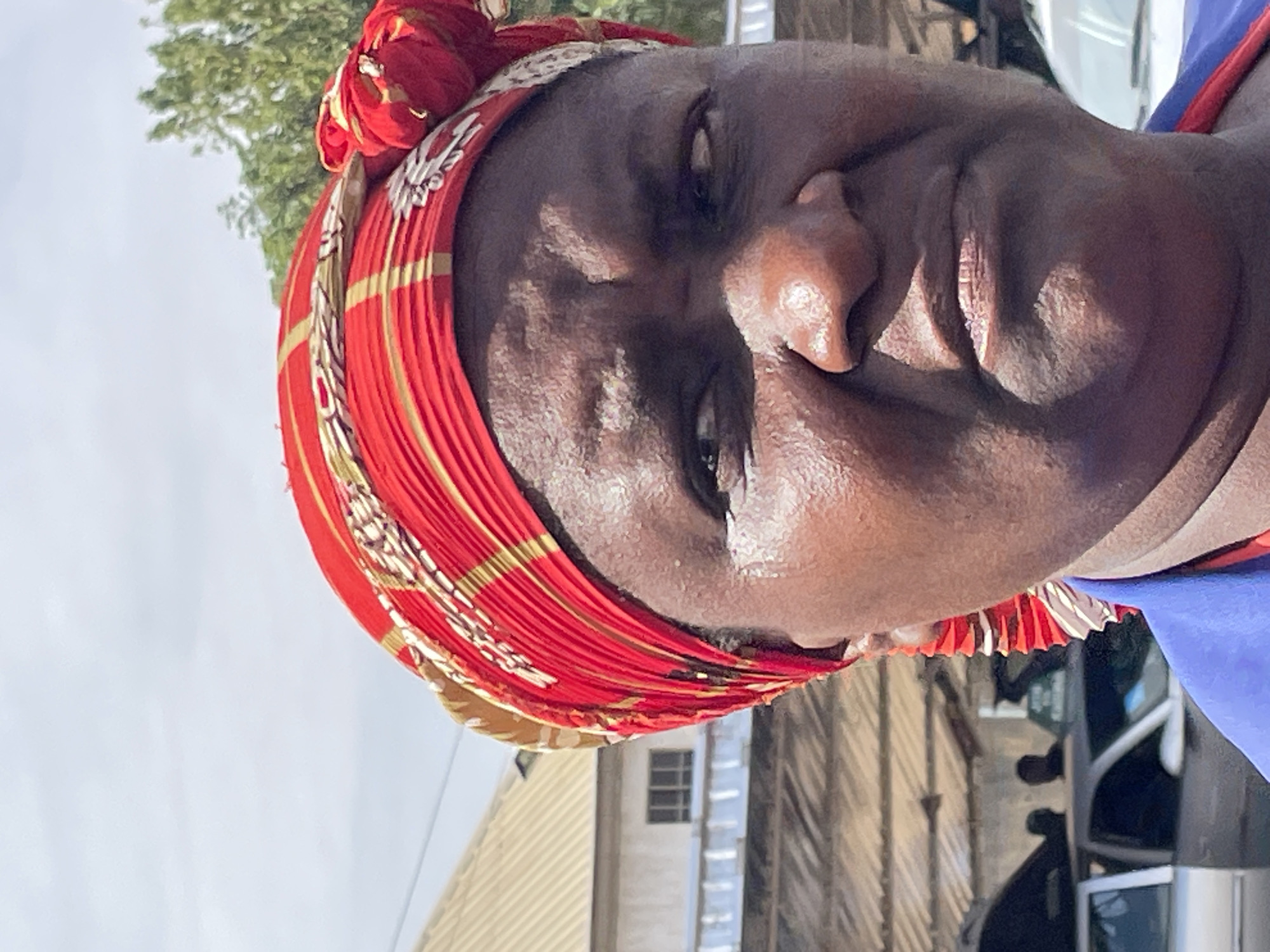 A close-up, rotated portrait of a dark-skinned woman with a red, gold, and white patterned headwrap. The background shows a building with a small window, a partly visible vehicle, and a tree. The location is inferred to be in Bukuru, Nigeria, based on the prompt instructions.