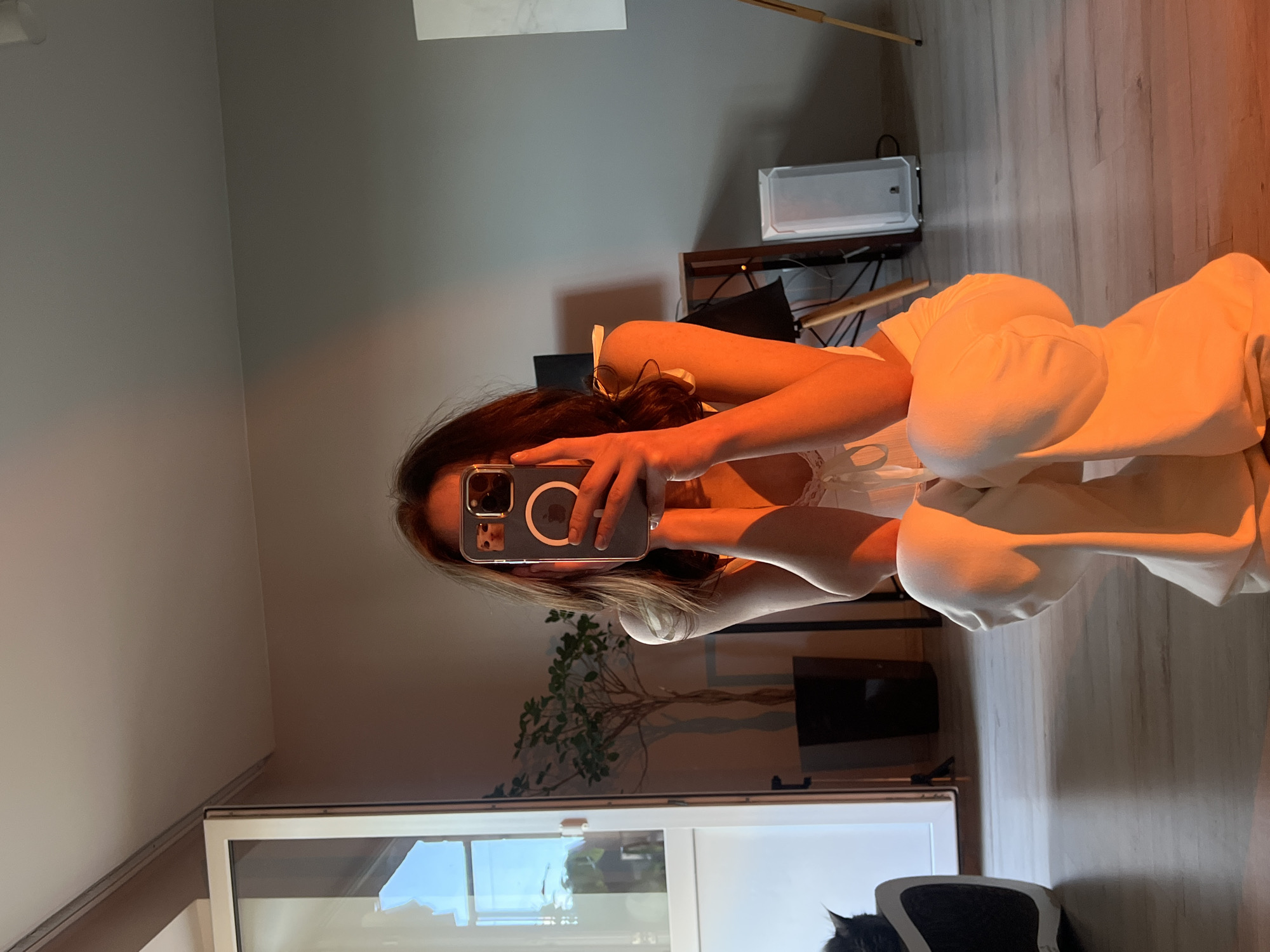A person, with long dark hair and fair skin, is crouching in a room, taking a selfie with their phone. They are wearing a white camisole and loose-fitting white pants. The room has light-colored wooden flooring and grey walls. To the left, there is a glass door, through which a potted plant and a black object, possibly a pet, are visible. In the background, there are a few pieces of furniture, including a shelf with items, a white rectangular object, and an easel. The scene is illuminated by warm, orange-toned light, suggesting it might be sunset or artificial lighting. No information about the location in Ternopil, Ukraine is present in the image.