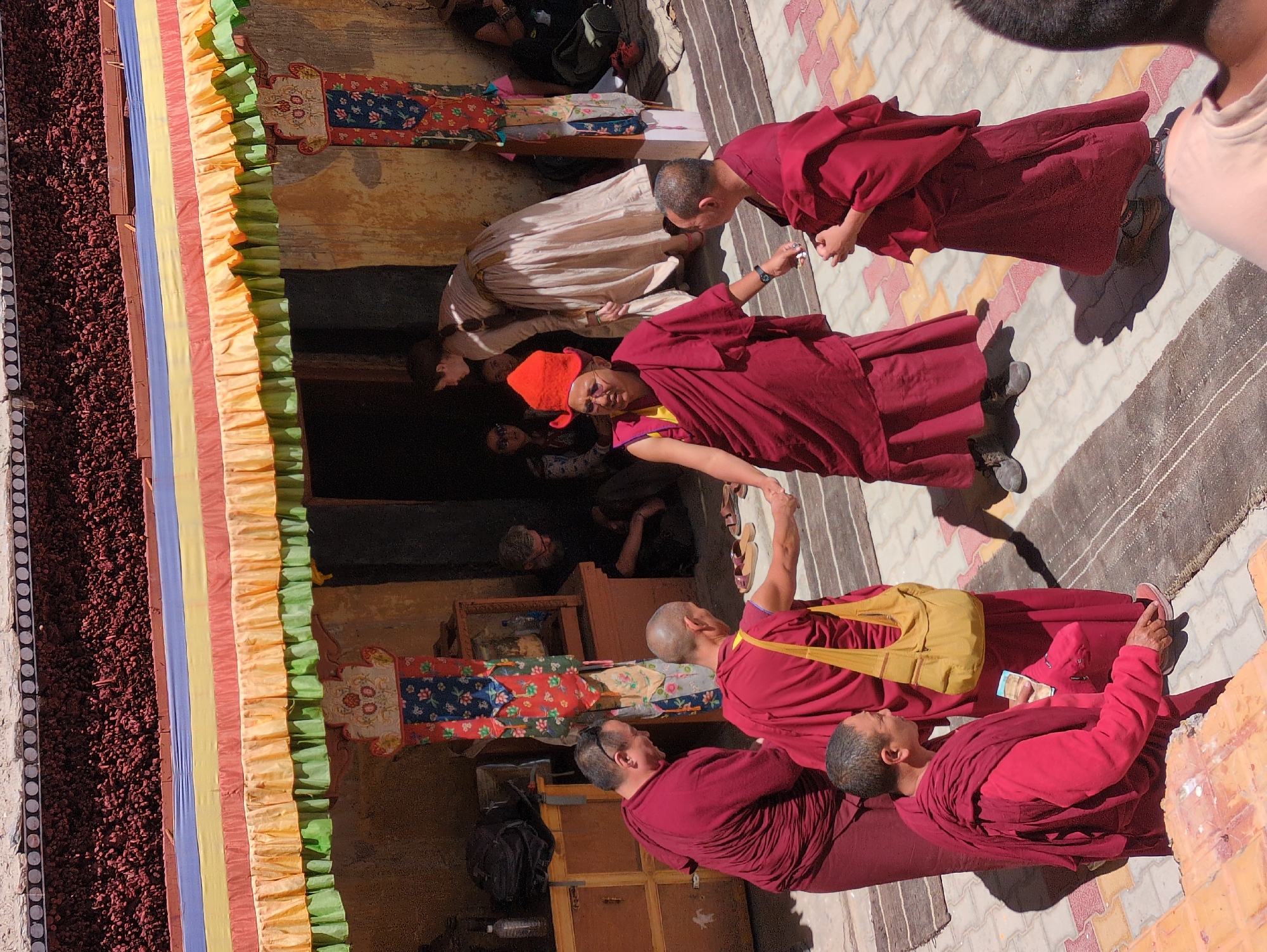 In Pibiting, India, a group of Buddhist monks and visitors are gathered outside a building adorned with colorful, striped awnings. Several monks in crimson robes are the central figures. One monk, wearing an orange conical hat and glasses, stands facing forward, holding a hand out. Another monk, with a yellow bag slung over his shoulder, is shaking hands with a third monk who is also wearing a crimson robe. In the background, a woman in a light-colored, long dress is seen entering a doorway, and other individuals are visible in the shadows. The ground is paved with a mosaic of colored tiles and striped rugs. The building features decorative pillars and a textured, dark roofline.