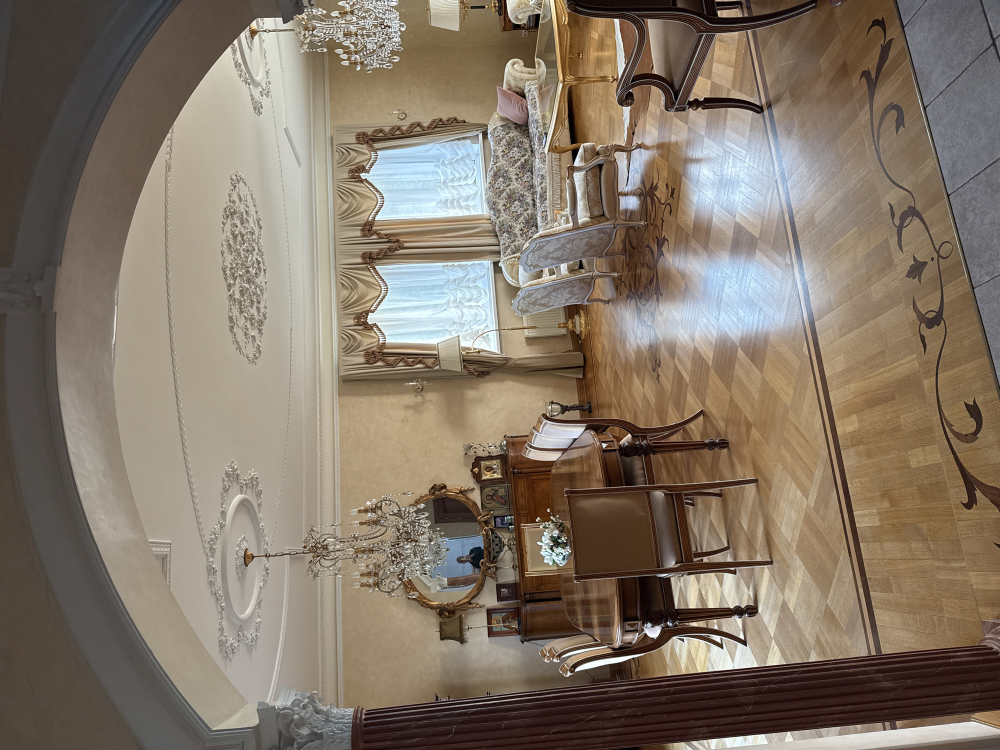 The image shows an opulent dining and living area in a home in Yukki, Russia. The room features ornate chandeliers, plush furniture, and intricate ceiling moldings. A large wooden dining table with six chairs is in the foreground, set for a meal. In the background, a floral-patterned sofa and two armchairs are arranged around a glass coffee table. The room is decorated with religious icons and a large ornate mirror above a sideboard. The flooring is a polished wood parquet with decorative inlays. The overall impression is one of wealth and traditional elegance.
