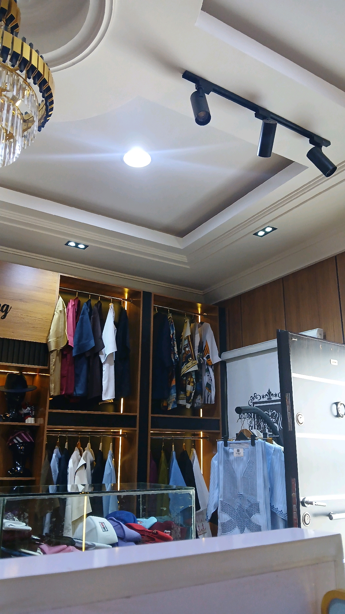 The image shows the interior of a clothing store. A chandelier hangs from the ceiling, and track lighting illuminates the merchandise. Racks of clothing, including shirts and traditional Nigerian attire, are displayed on wooden shelves. In the foreground, a glass display case contains hats and other accessories. The store appears to be well-lit and organized, with a modern and luxurious aesthetic.