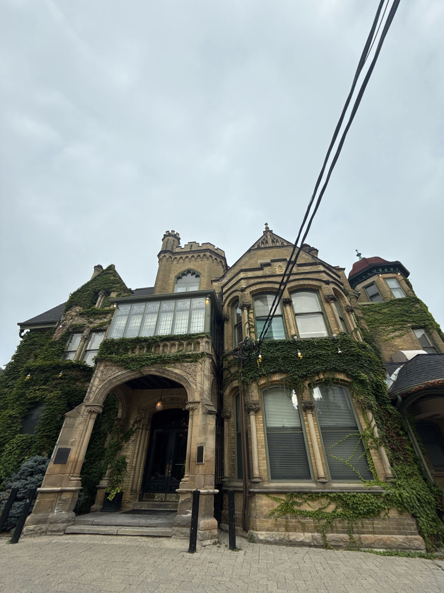 This is an eye-level, ground-level exterior, low-angle shot of a large, old, ivy-covered building that appears to be in the Annex neighborhood of Toronto, Canada. The building is constructed of yellow brick and features Gothic architectural elements, including arched doorways, turrets, and ornate window frames. The ivy covers a significant portion of the building's facade, giving it a historic and slightly overgrown appearance. The sky is overcast and a utility pole with three black wires is visible in the upper right portion of the frame. The entrance to the building is under a stone archway, with a dark wooden door and a stone mat in front. The foreground shows a cobblestone walkway leading up to the entrance. There are no people visible in the image.