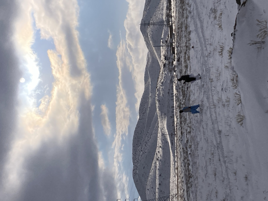 The image depicts a snow-covered landscape in Churmian, Pakistan, under a partially cloudy sky with sunlight breaking through a circular opening in the clouds in the upper-right quadrant. The foreground consists of a vast, flat expanse of snow with sparse, dry vegetation protruding, and faint tracks are visible on its surface. Two individuals are walking from left to right across this snow-covered field. The person on the left wears light blue, loose-fitting trousers and a dark upper garment. The person on the right is dressed in dark clothing and appears to be holding a dark, rectangular object, possibly a mobile phone, near their chest. In the mid-ground and background, undulating hills are extensively covered in snow, with some darker patches of exposed ground or vegetation. Tall lattice transmission towers are present on both the far left and far right sides of the image, extending into the mid-ground. A small cluster of buildings or structures is visible in the distant mid-ground, positioned at the base of the snow-covered hills on the right side.