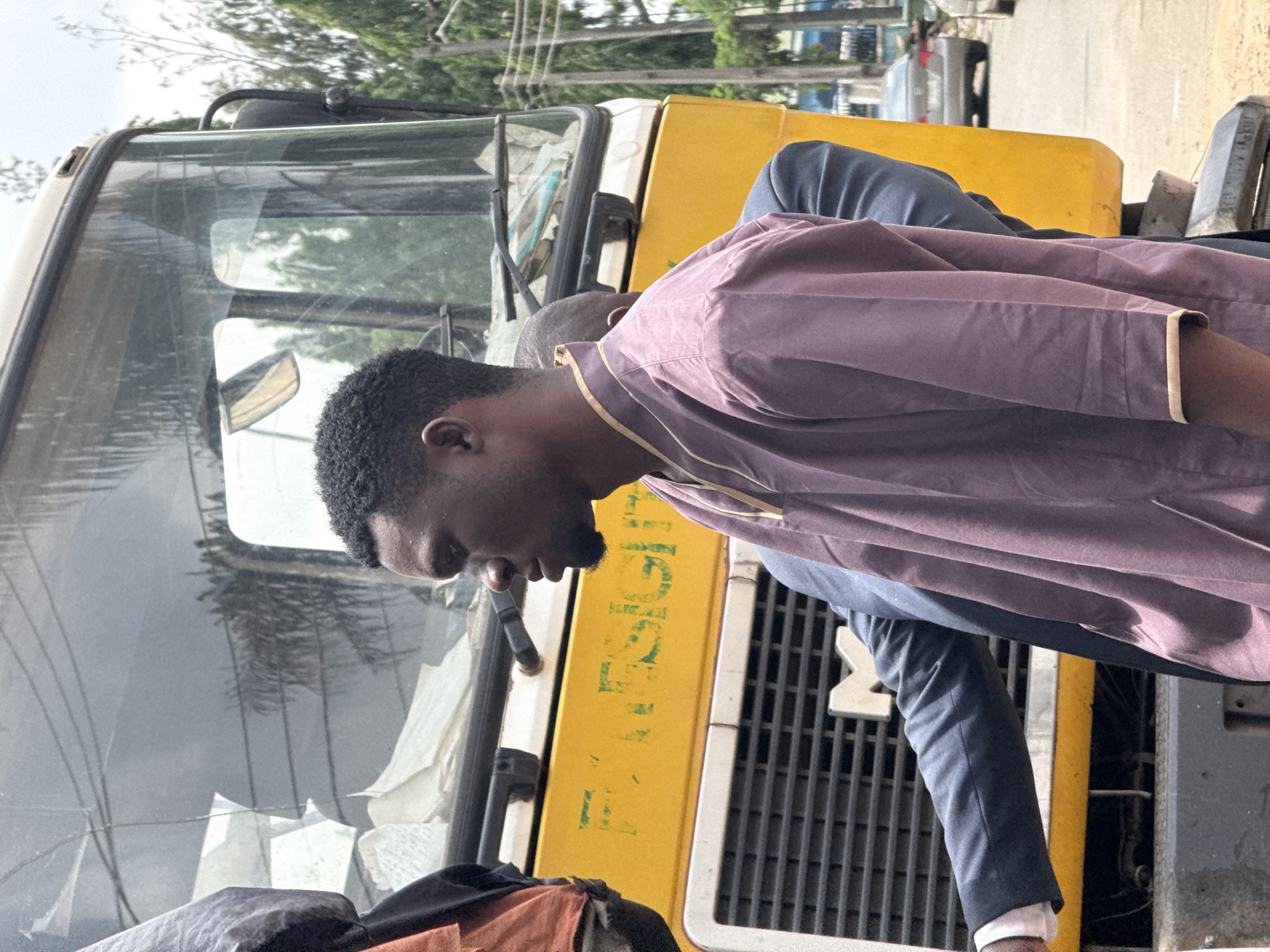 In Abuja, Nigeria, a medium shot captures a young Black man from the chest up, facing right in profile. He has short, dark, curly hair and a light beard. He wears a purple garment with gold trim at the neckline and cuffs. He is standing in front of a large, yellow truck. Behind him, partially obscured, is another person wearing a dark suit jacket and a light-colored shirt cuff. The truck has a large, grimy windshield with wipers. White paper or fabric is visible inside the cab. The front of the truck is yellow with a silver grille featuring the letter "M". To the right of the truck, a grey sedan is visible on a paved road, with trees and utility poles in the background. The overall impression is an outdoor, urban setting.