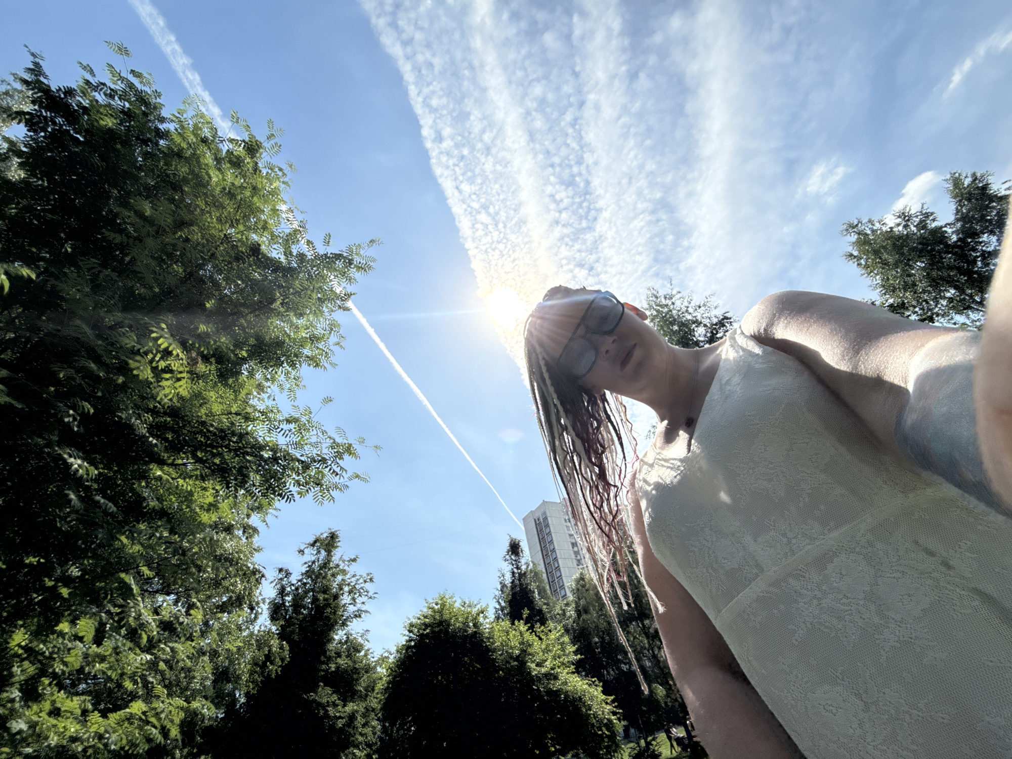 This image depicts an outdoor scene captured from a low-angle perspective, looking upwards into a bright sky, characteristic of a park within a residential area in Moscow, Russia.
In the foreground, a female-presenting individual is partially visible, primarily in the lower right quadrant. They are wearing dark sunglasses that obscure their eyes and have numerous long, multi-toned dreadlocks or braids. The individual is dressed in a light-colored, likely off-white, sleeveless dress with a distinct lace-like floral pattern. A faint blue or greenish tattoo is visible on their right upper arm. Their head is tilted significantly backward, indicating they are looking down towards the camera, which is positioned very low.
The upper portion of the image features a vivid blue sky, intensely bright at the center due to the sun, which causes a significant lens flare emanating from the upper-center. High-altitude, wispy cirrus clouds form distinctive patterns across the sky, particularly a textured, rippled band in the upper right. A prominent, straight white contrail from an aircraft cuts diagonally across the sky from the upper left to the mid-right.
Dense green foliage from several large deciduous trees occupies the left and lower left sections of the frame, with sunlight dappling through their leaves. In the mid-ground, partially obscured by trees and against the sky, stands a tall, multi-story residential building. This building exhibits the typical architectural style of a Soviet-era apartment block, characterized by its numerous windows and concrete or light-colored facade, common throughout Moscow. A thin wire or power line is faintly visible stretching across the sky near the building. The overall setting is consistent with a public park or green space adjacent to urban residential development in Moscow, Russia.