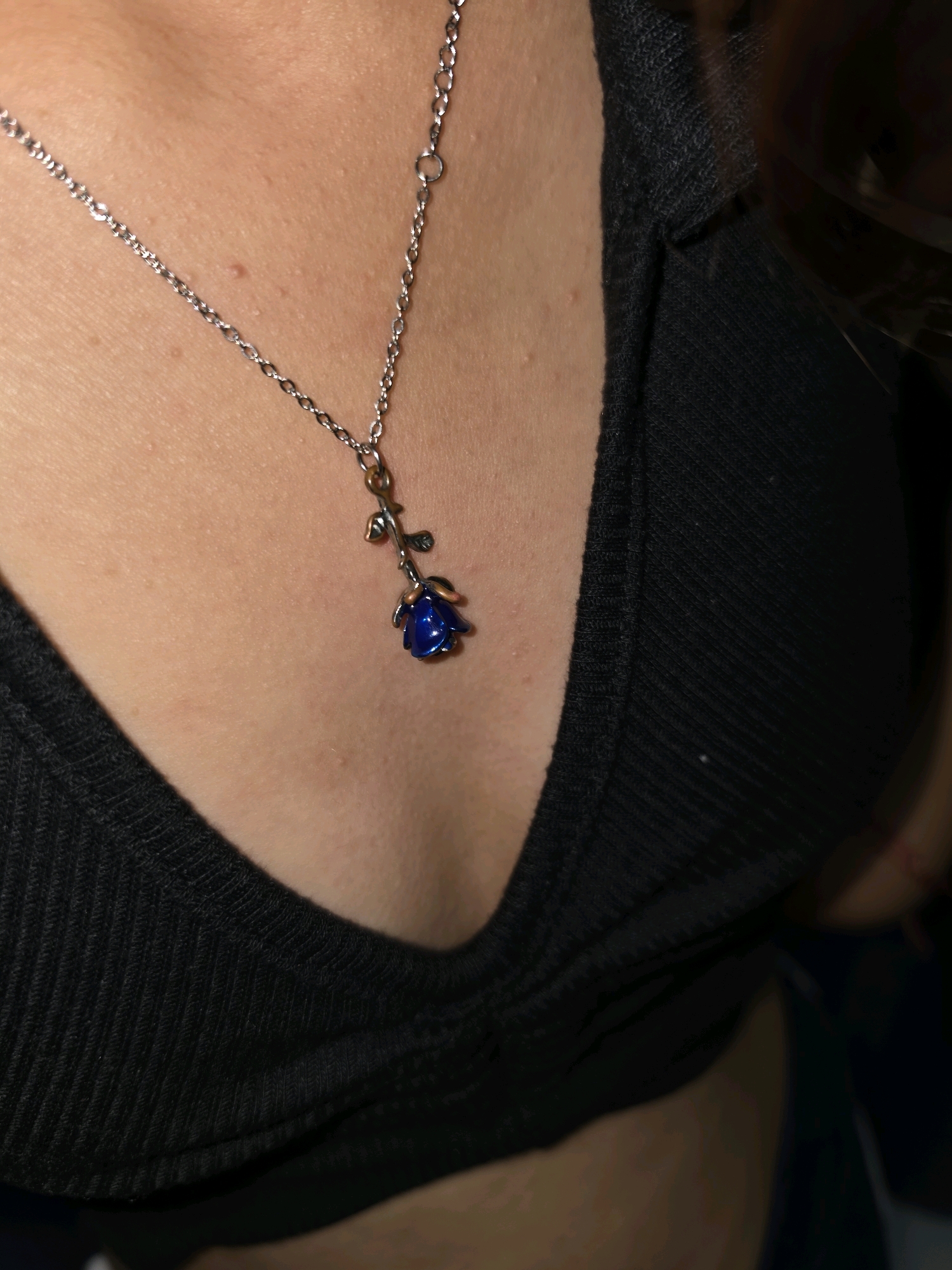 The image shows a close-up of a person's chest wearing a black ribbed top and a silver necklace with a small blue rose pendant. The necklace chain is thin and delicate, with a clasp visible near the neckline. The pendant is shaped like a rose bud, with detailed petals in a vibrant blue color and a silver stem and leaves. The skin is visible around the necklace, showing a few minor blemishes. The lighting is dim, creating shadows that highlight the textures of the clothing and skin. The background is dark and out of focus, obscuring any environmental details. The image is taken from a high angle, looking down at the person's chest.