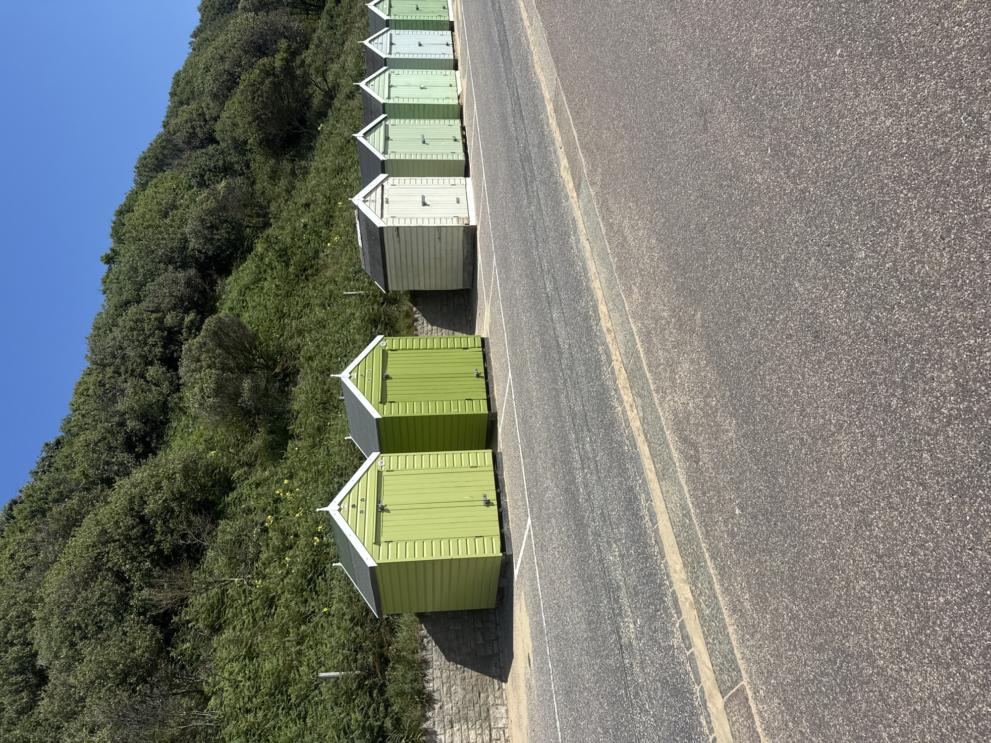 The image shows a row of beach huts along a promenade in Bournemouth, United Kingdom. The huts are painted in various shades of green and white, with traditional pitched roofs. They are situated on a paved area next to a road and a grassy slope covered in dense foliage. The sky is clear and blue, indicating a sunny day. The scene is quiet and appears to be unoccupied. The huts are lined up neatly, suggesting they are part of a rental or private collection. The overall impression is one of a picturesque coastal setting.