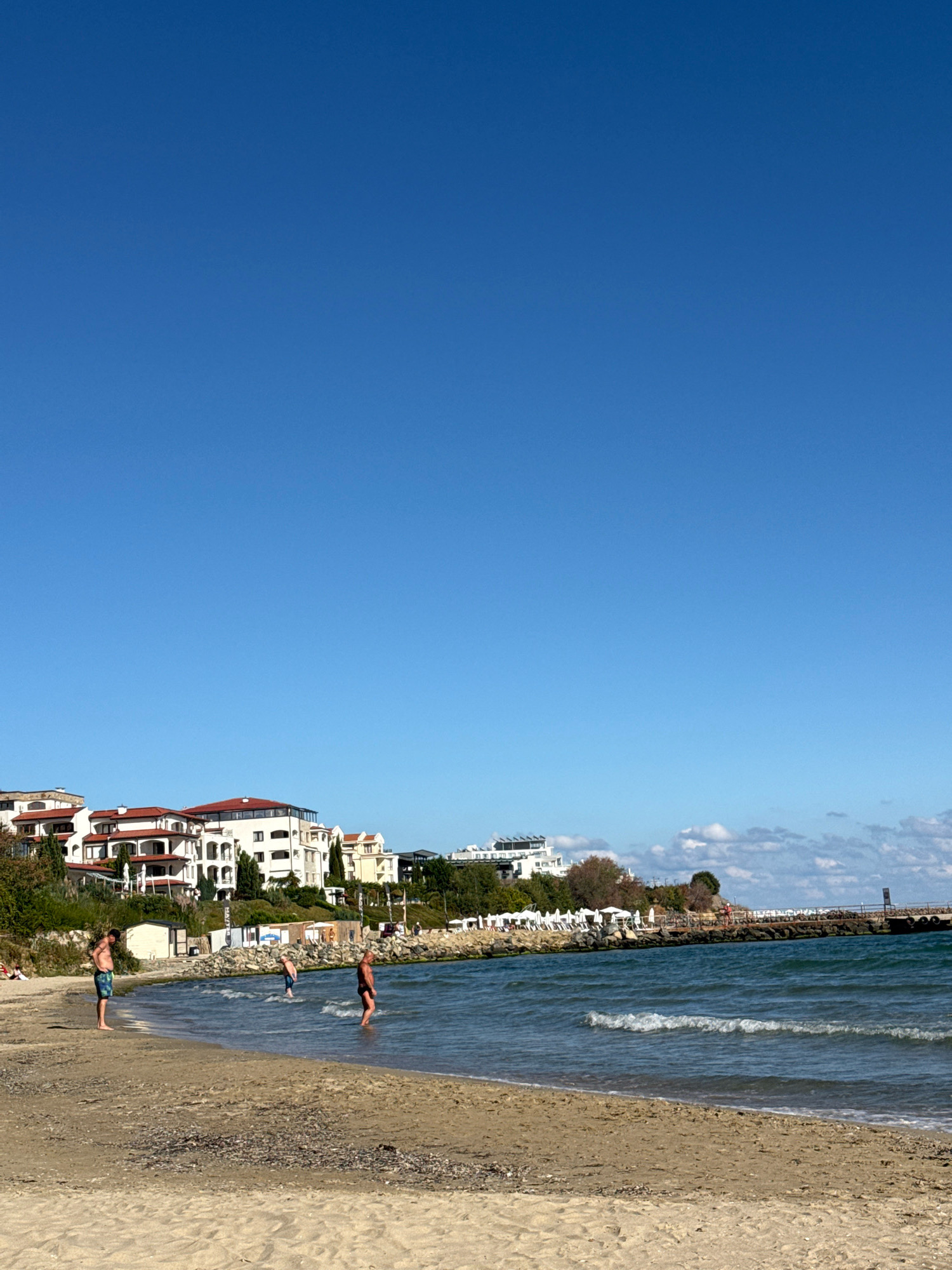 The image depicts a beach scene in Sveti Vlas, Bulgaria, on a clear day with a vibrant blue sky. The foreground is dominated by a sandy beach with scattered debris. In the mid-ground, the turquoise water laps gently onto the shore, with small waves visible. Three individuals are in or near the water. To the left, a man stands on the wet sand, facing away from the camera. Further into the water, another man stands, partially submerged. To the right, a third man stands in the water. The background features a coastal area with a line of white buildings with red-tiled roofs, characteristic of Mediterranean architecture, situated on a gentle slope. Some trees and greenery are interspersed among the buildings. A rocky breakwater or pier extends into the sea from the right side of the image, with a navigational marker at its end. There are also beach chairs and umbrellas visible near the buildings. The overall scene conveys a sense of a relaxed summer day at the seaside.