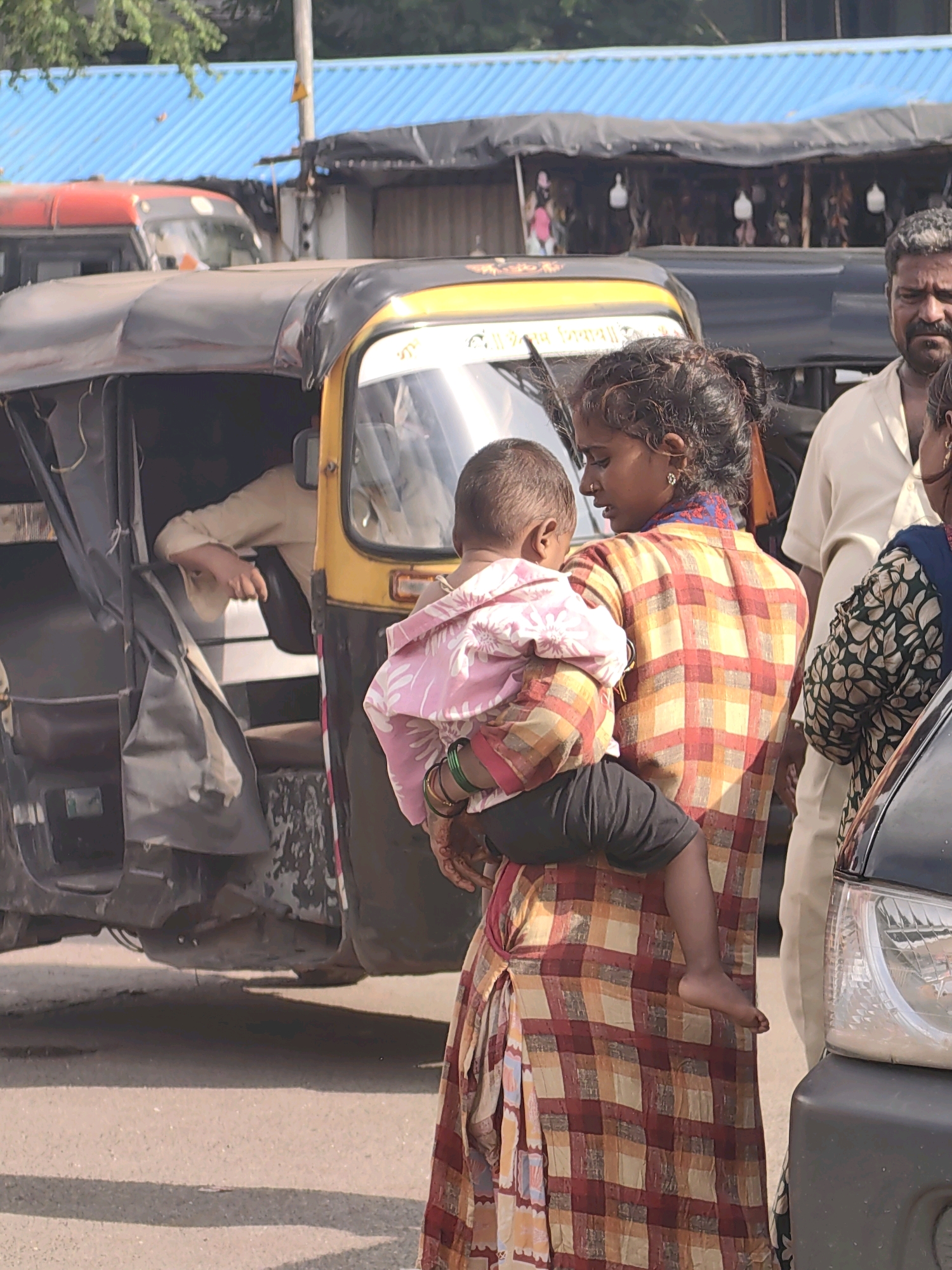 A woman in a checkered dress is holding a baby wrapped in a pink blanket. They are standing near an auto-rickshaw. A man in a white shirt stands behind them, and another person wearing a patterned top is to the right. In the background, there are stalls with blue awnings and a red vehicle. The scene appears to be on a street in Mumbai, India.