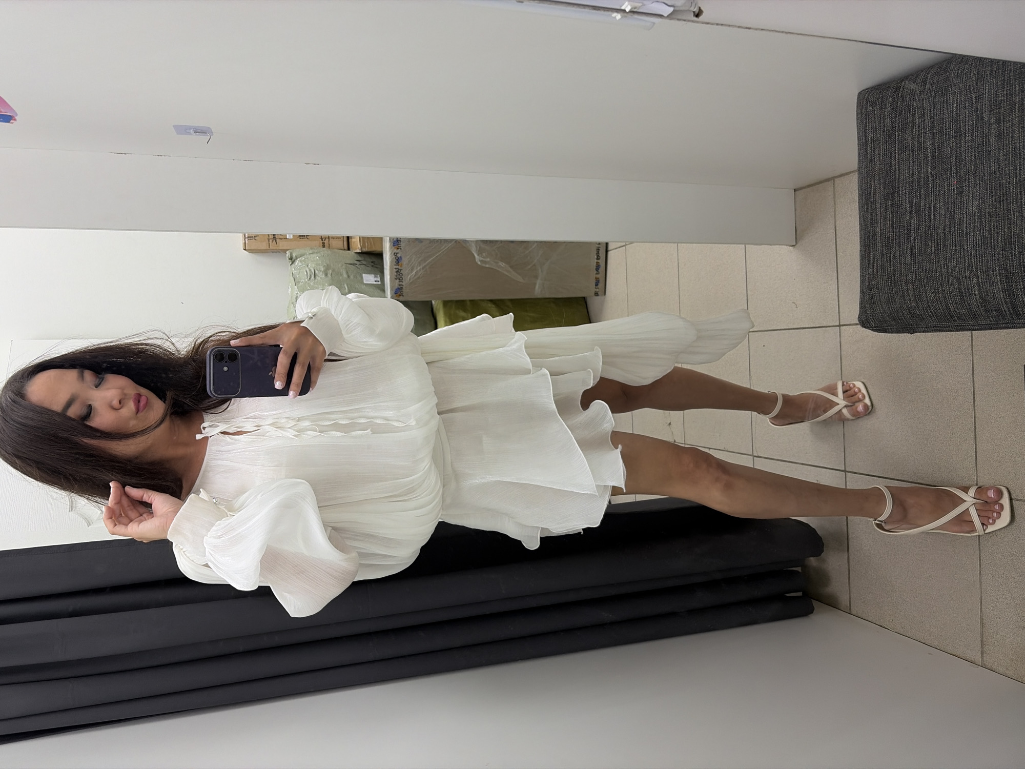 An Asian woman with dark hair is standing in a changing room, taking a mirror selfie. She is wearing a white, flowy dress with puffed sleeves and a layered skirt, paired with white heeled sandals. The changing room has a dark curtain on the left and white walls on the right. In the background, there are boxes wrapped in plastic. The floor is tiled. The woman is holding a purple smartphone in her right hand and touching her hair with her left hand.