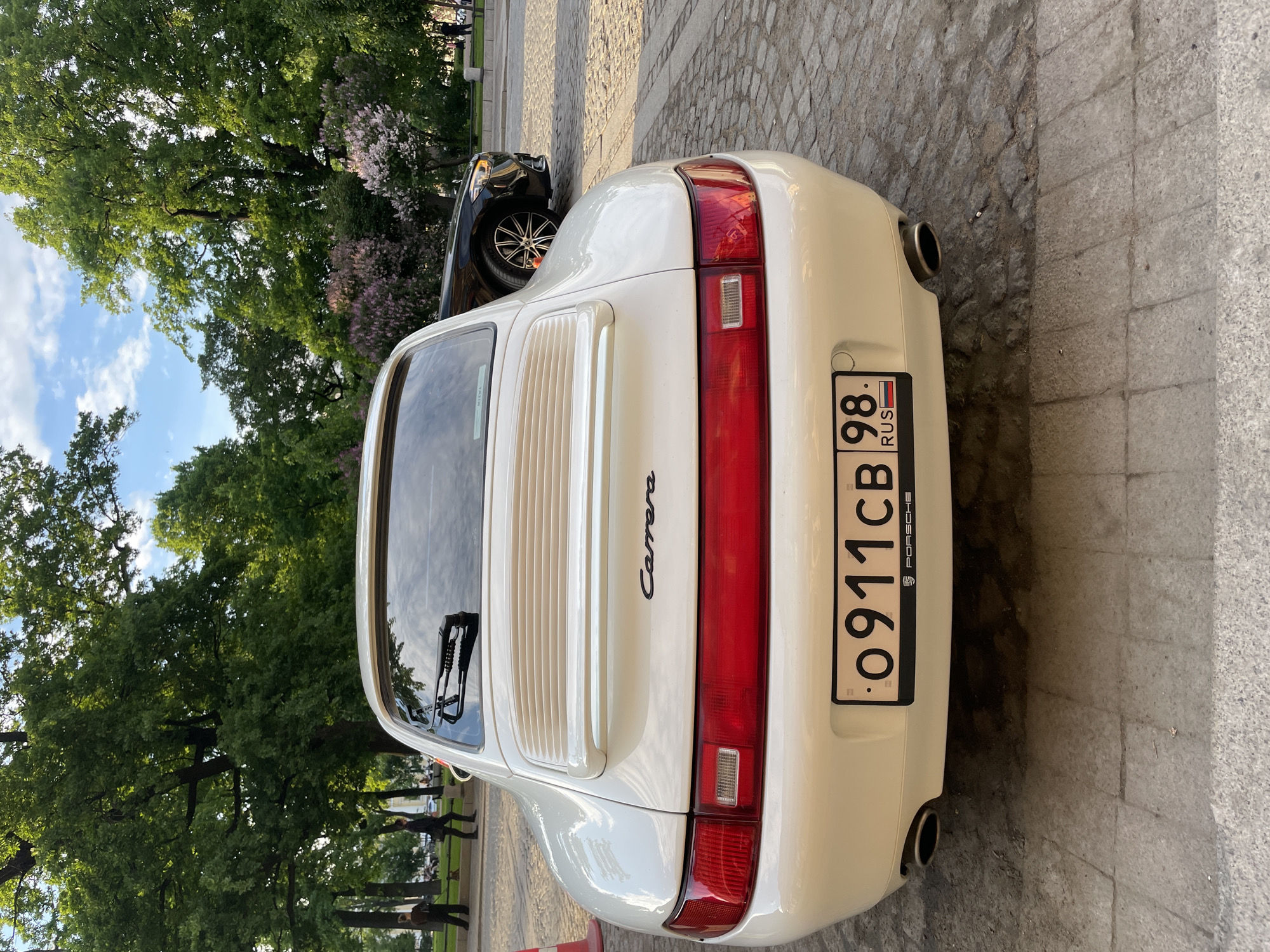 An overhead, slightly angled rear view captures a white Porsche 911 Carrera parked on a paved surface in Saint Petersburg, Russia, under bright daylight. The scene suggests a pleasant day, with a clear blue sky and fluffy white clouds visible through lush green trees and flowering lilac bushes in the background.
The primary subject is the white Porsche, an older generation 911, showcasing its classic design. The car features a prominent red full-width taillight bar spanning the rear, with distinct rectangular reverse lights/indicators on either side. Its engine lid is characterized by horizontal slats and bears the "Carrera" script emblem. A single rear wiper is visible on the back window. Dual round exhaust pipes protrude from beneath the rear bumper.
The Russian license plate "O 911 CB 98 RUS" is clearly visible, with "98" denoting the Saint Petersburg region, and the word "PORSCHE" printed on the plate frame.
Behind the Porsche, partially obscured, is a black SUV with multi-spoke alloy wheels. The ground consists of uneven grey cobblestones transitioning into smoother stone or concrete slabs. In the distant background, several indistinct figures of people can be seen walking or standing on a path amidst the trees, suggesting a public park or street setting. A small red and white traffic cone or barrier is barely visible on the ground near the Porsche's rear passenger side.