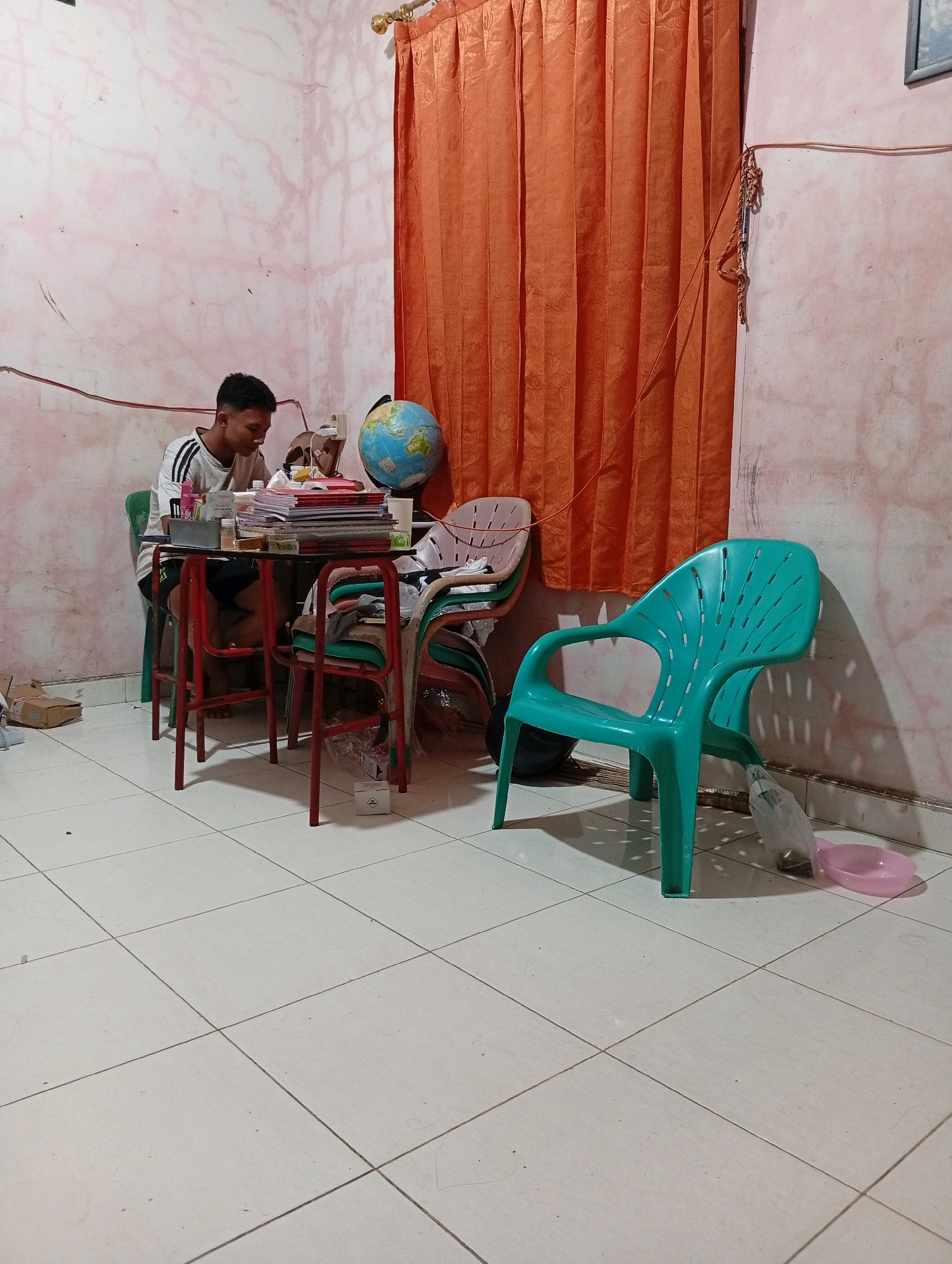 In the city of Gunungsitoli, Indonesia, a young man sits at a table, engrossed in his studies. The room is lit by an unseen light source, casting shadows on the pale pink, slightly textured walls. An orange curtain hangs on the right side of the frame, partially obscuring a window. On the table, a stack of books and papers are spread out, alongside a globe and a bottle of pink liquid. The young man, wearing a white t-shirt with black stripes on the sleeves, is seated on a green chair. Several other chairs are present in the room, including a teal-colored plastic chair in the foreground, and other chairs with red metal frames. The floor is tiled with white square tiles. On the floor near the teal chair, a clear plastic bag with some items inside and a pink basin are visible. The overall impression is that of a humble study space in a residential setting.