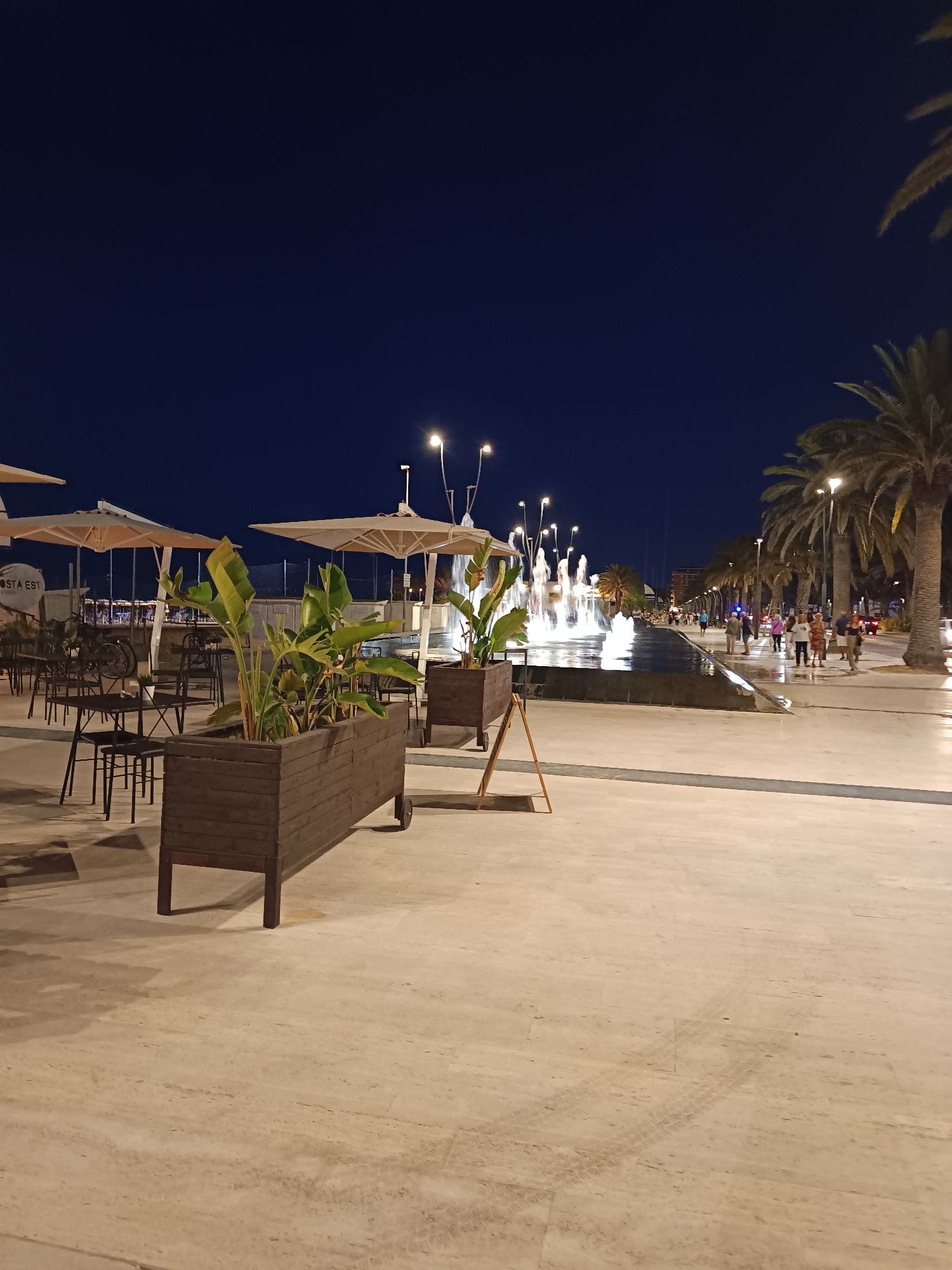 The image depicts a night scene in Roseto degli Abruzzi, Italy, likely along the Lungomare. In the foreground, there is a paved walkway with several outdoor tables and chairs from what appears to be a restaurant or cafe. Large, beige umbrellas are set up over the seating area. A prominent wooden planter box filled with lush green plants is positioned in the mid-ground, adding a touch of greenery to the scene.
Further into the image, a decorative water fountain with multiple jets of illuminated water is active, creating a dynamic focal point. Beyond the fountain, a tree-lined promenade stretches into the distance, with palm trees lining the right side of the walkway. Several people are seen strolling along the promenade, engaged in evening activity. Streetlights and building lights are visible in the background, illuminating the urban environment. The sky is dark, indicating it is nighttime. The overall atmosphere suggests a lively public space designed for evening recreation and dining.