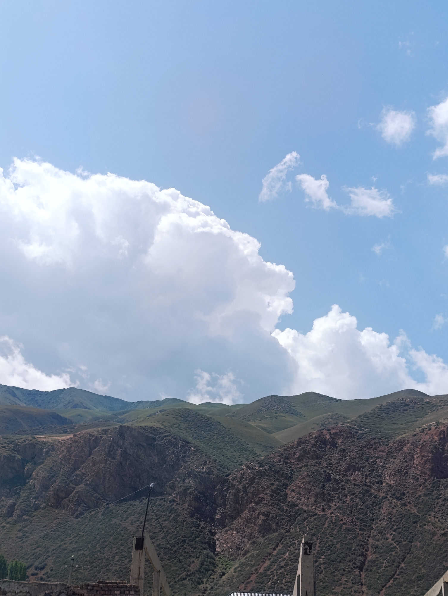 A high-angle, full shot captures a mountain range under a bright, partly cloudy sky. The mountains are covered in green vegetation and some rocky, reddish-brown soil. The sky is predominantly blue with large, fluffy white clouds in the upper left and smaller scattered clouds throughout. In the foreground, partially visible structures made of concrete and brick suggest some form of construction or development. A black cable stretches across the lower portion of the image, connected to a pole. The scene appears to be an outdoor environment, possibly in the outskirts of Naryn City, Kyrgyzstan, given the mountainous terrain and architecture.