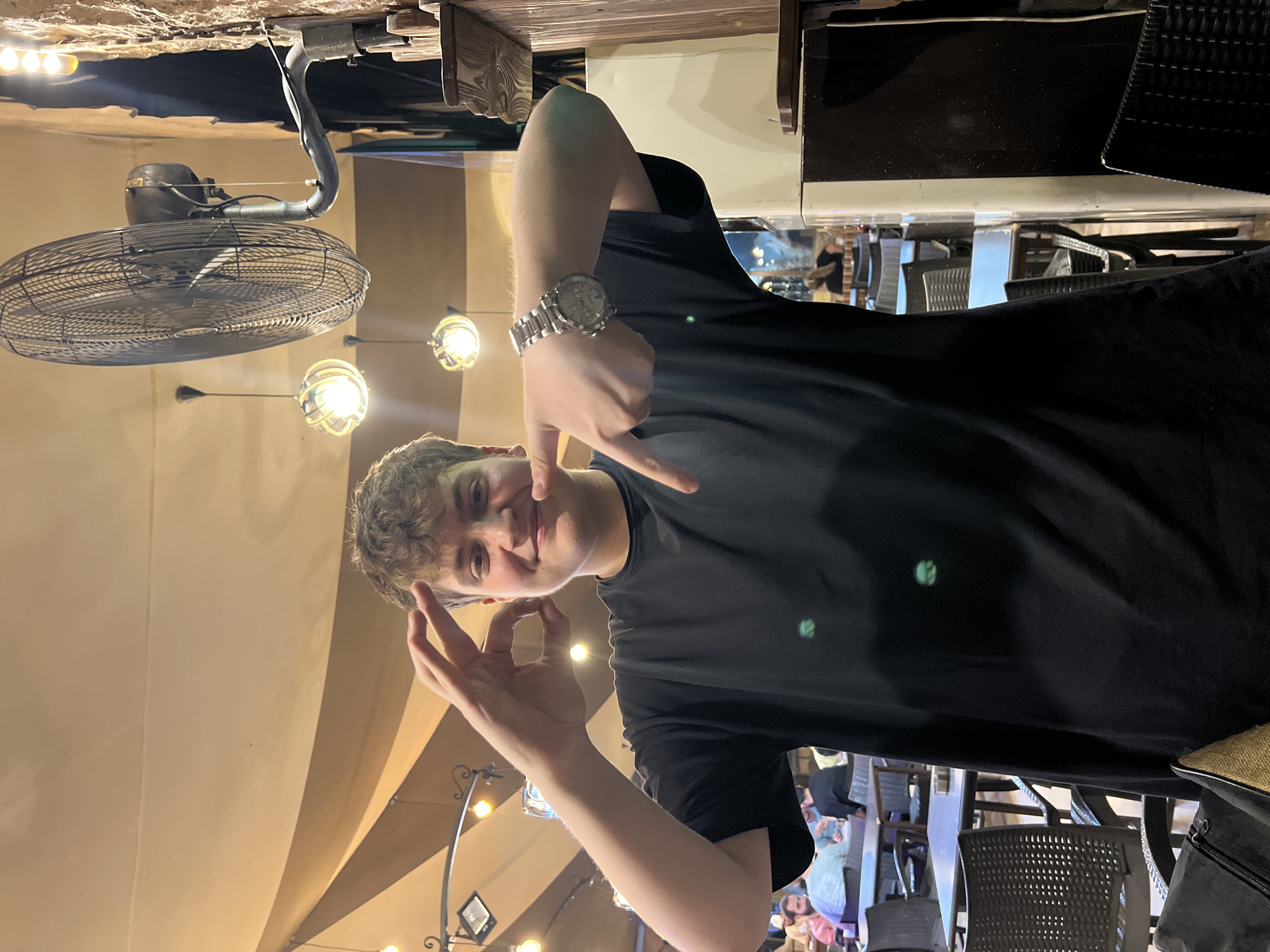 A young man is captured in a close-up, waist-up shot, posing inside what appears to be a restaurant or cafe in Rukn ad-Din Municipality, Syria. He has light brown, curly hair and is wearing a plain black t-shirt and a silver-toned watch on his left wrist. The man is smiling broadly at the camera, making distinct hand gestures: his right hand points towards his face, while his left hand forms an "OK" sign.
The indoor setting features a distinctive ceiling structure with light-colored draped fabric and dark wooden beams. Several artificial light sources are visible, including ornate spherical lights encased in metal cages and smaller recessed LED lights. A large, dark industrial-style ceiling fan hangs prominently in the upper left portion of the image. In the blurred background, other people are visible seated at tables and chairs, suggesting a lively dining atmosphere. A black bag or backpack is partially visible on the far right.
The artificial lighting implies the photograph was taken in the evening or at night. The subject's expression conveys happiness and a playful mood. No discernible text is visible within the image.