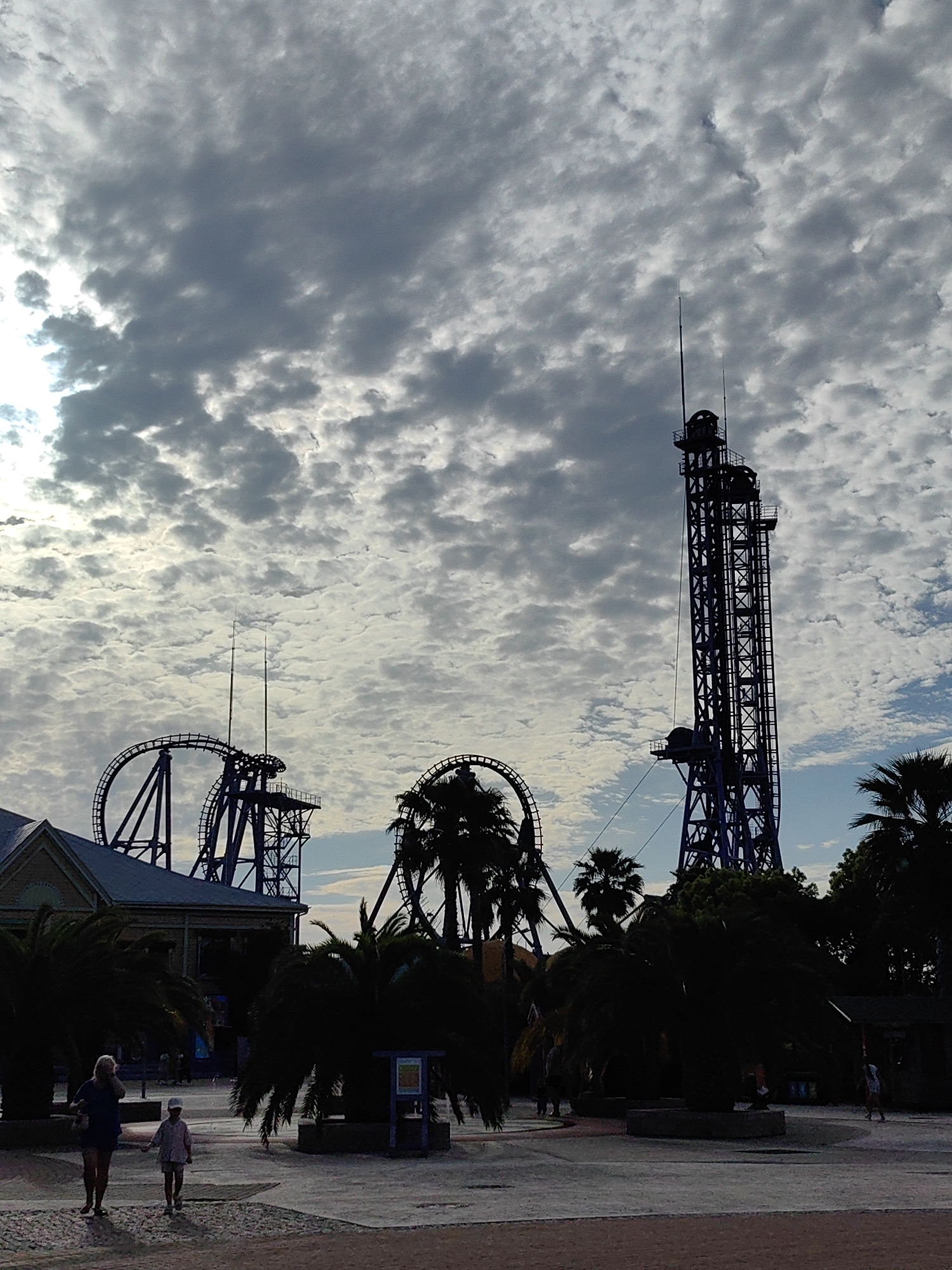 The image depicts an amusement park in Sirius, Russia, under a cloudy sky. A tall, dark purple structure, likely a thrill ride like a drop tower, dominates the right side of the frame. To the left of this structure are sections of a roller coaster track, also in a dark purple hue. The roller coaster features a prominent loop and a steep drop.
In the foreground, the ground is paved with cobblestones and sand. Two people are walking on the paved area. A woman in a blue top and shorts walks towards the left, while a child in a light-colored shirt and shorts walks ahead of her, also towards the left. Several silhouetted palm trees are scattered throughout the scene, casting dark shadows. A building with a light-colored facade and a dark roof is visible on the left side of the image, behind which more roller coaster elements are present. A small kiosk or information booth is situated among the palm trees. The overall impression is that of an amusement park on a potentially overcast day, with the rides as the central features against the backdrop of the sky and tropical-like vegetation.