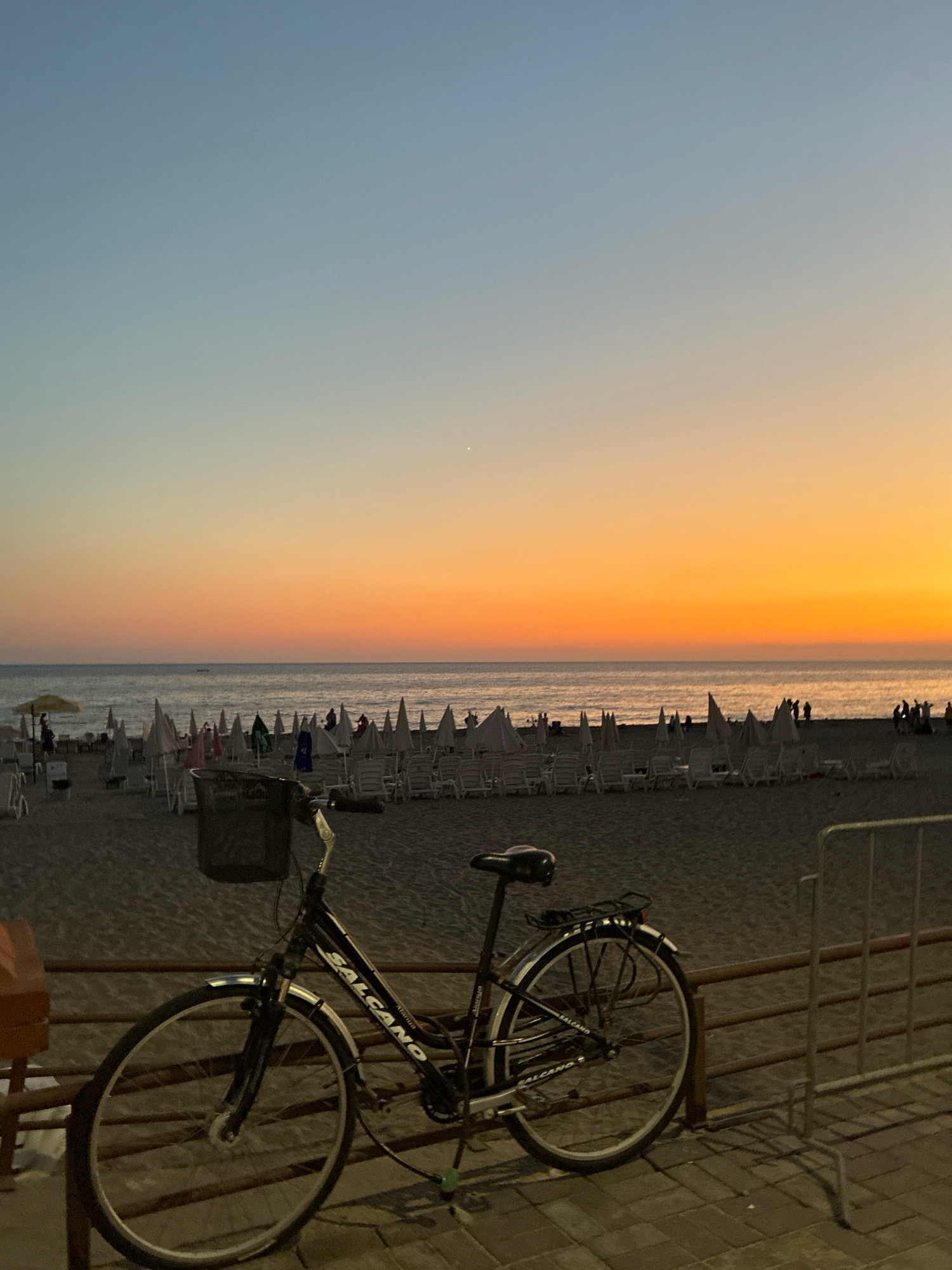 The image captures a serene evening at a beach in Alanya, Turkey, as evidenced by the "ALCANO" branding on the bicycle, a common Turkish bicycle brand. A bicycle with a basket is prominently displayed in the foreground, parked along a paved walkway. Beyond the walkway, a sandy beach stretches out, dotted with numerous white beach chairs and umbrellas, most of which are closed. In the distance, the calm sea meets the horizon under a gradient sky transitioning from a pale blue at the top to a vibrant orange and yellow sunset. A few figures can be seen walking on the beach, and a small boat is visible on the water. The scene is peaceful, suggesting the end of a day at the beach.